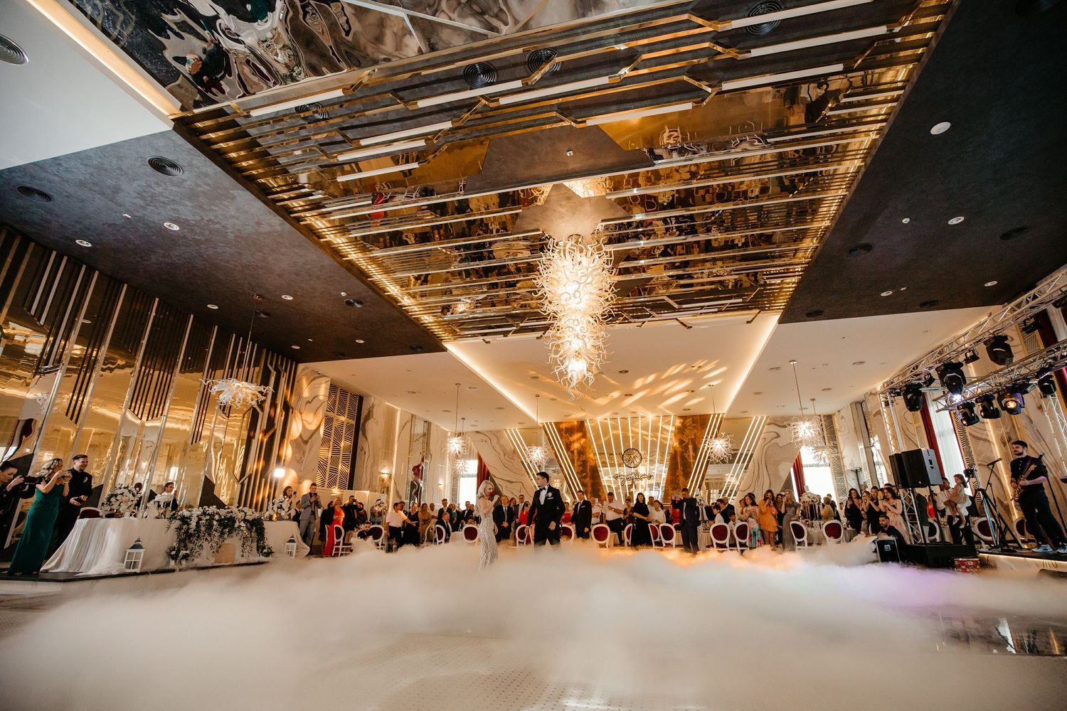 Grand wedding reception hall with mirrored gold ceiling, crystal chandeliers, and fog-covered dance floor.