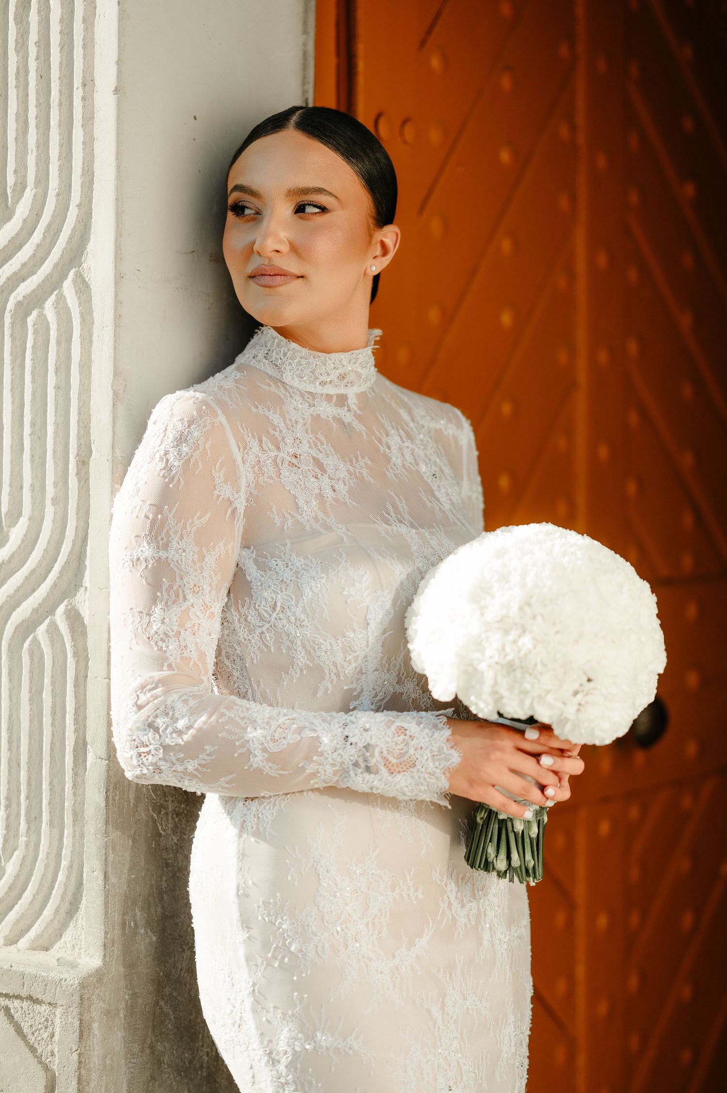 Elegant bride in long-sleeve lace wedding gown holding white bouquet, leaning against ornate door.
