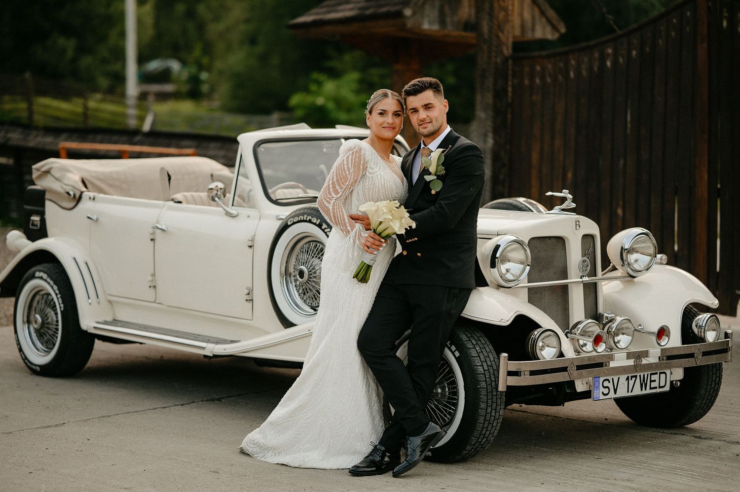 Elegant bride and groom posing beside a classic white vintage wedding car outdoors.