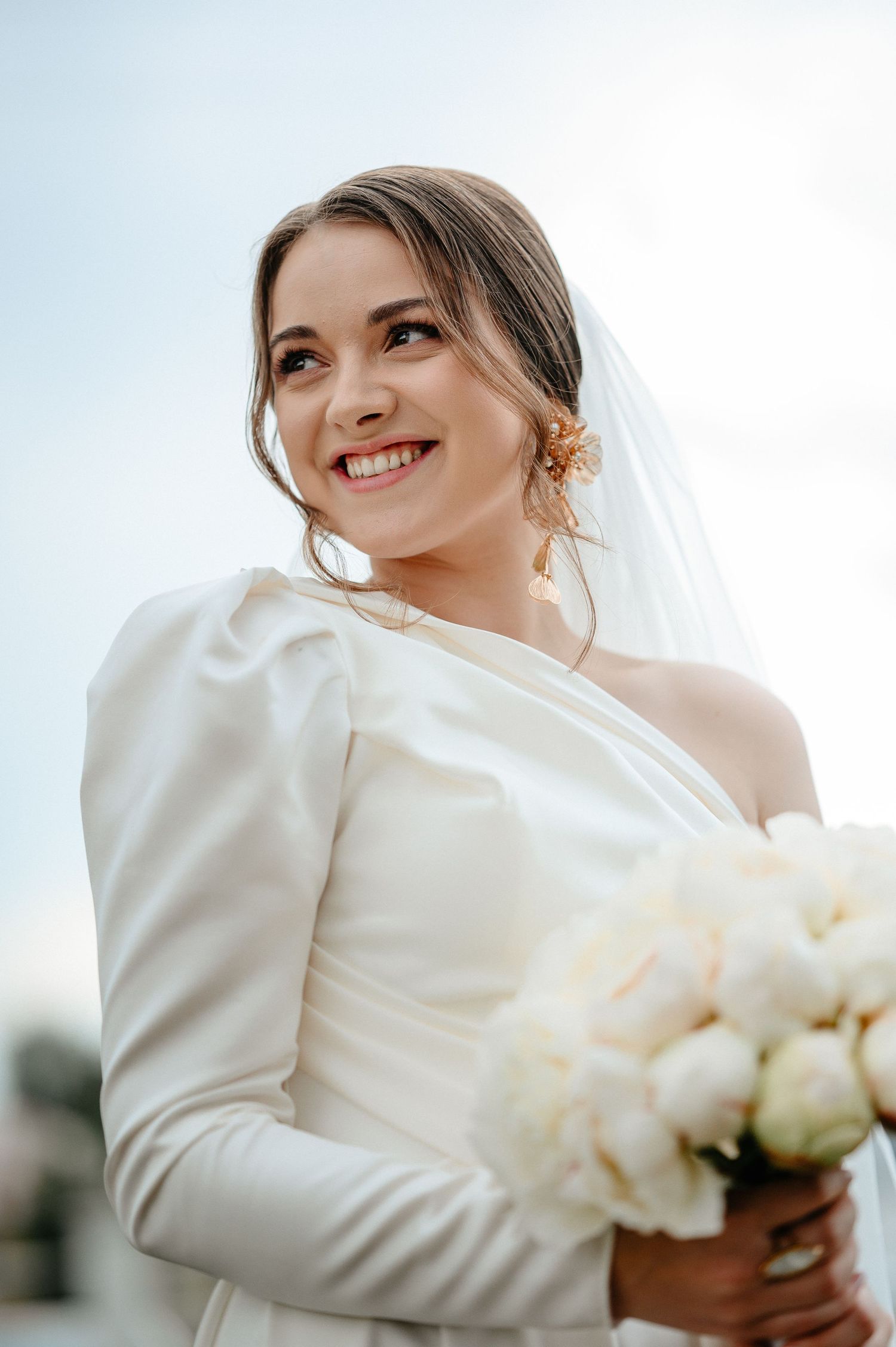 Radiant bride in elegant white one-shoulder gown holding peony bouquet under bright sky.