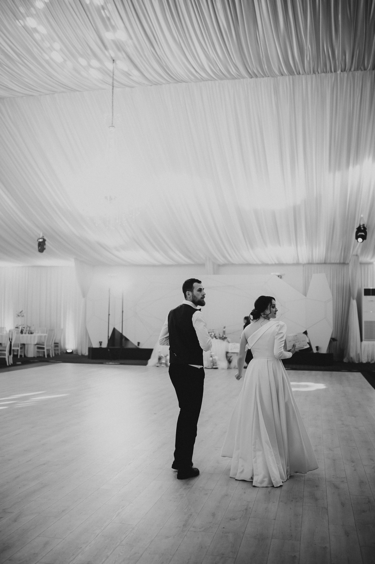 Black and white photo of bride and groom sharing first dance in elegant draped ballroom.