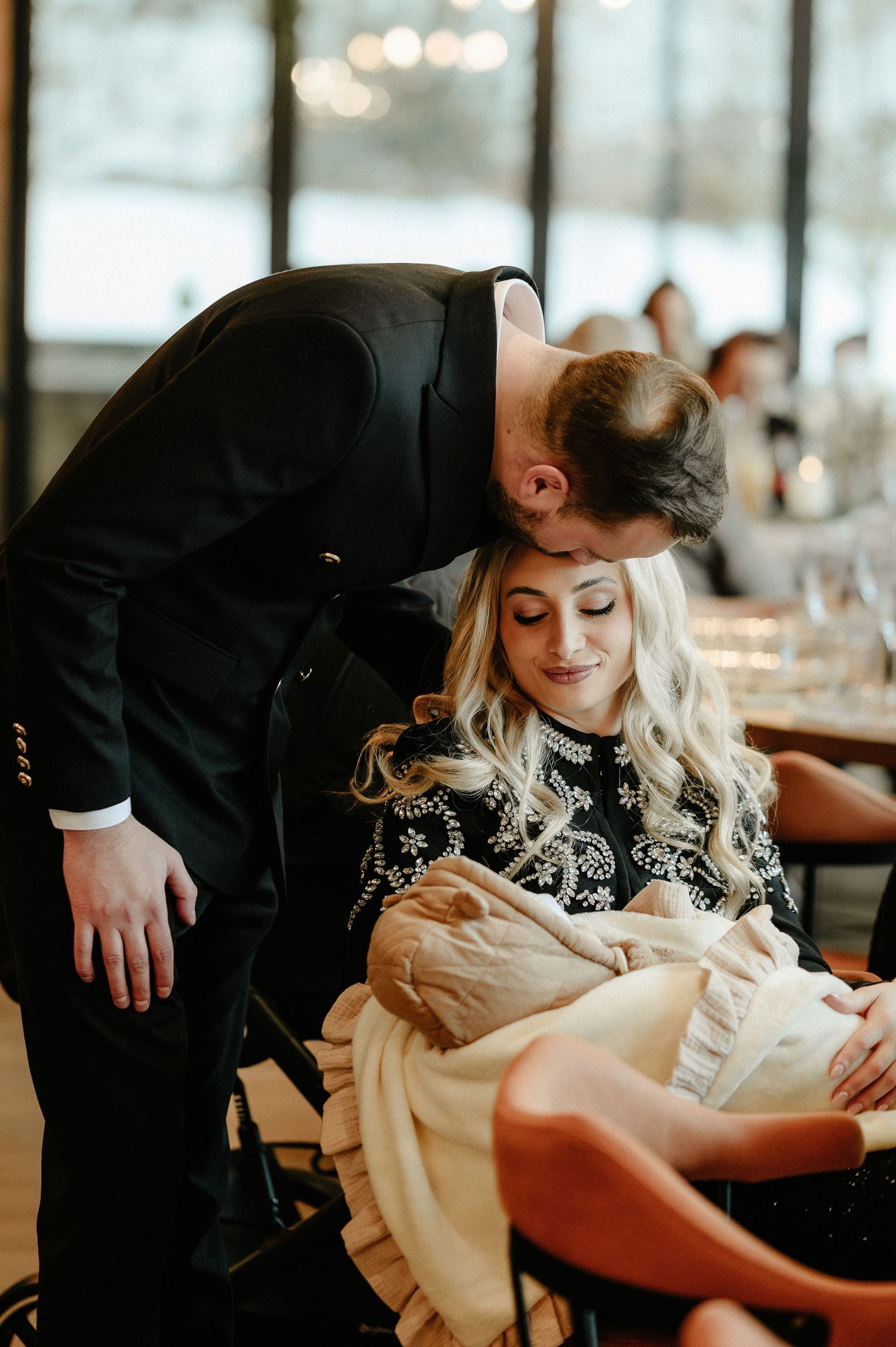 Tender moment as man in black suit kisses woman holding newborn baby at indoor reception.
