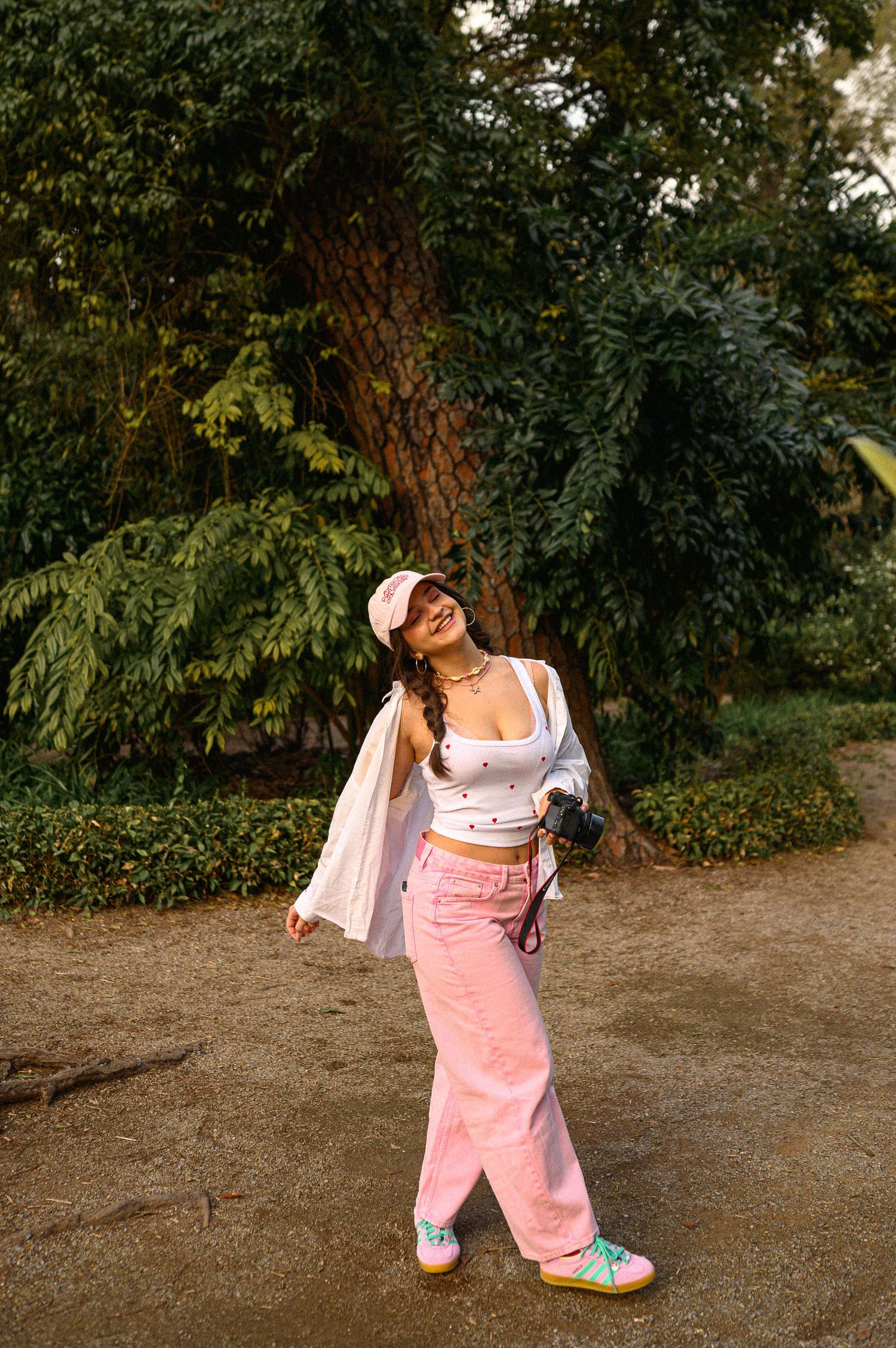 Stylish young woman in pink wide-leg pants and white top posing in a lush green park with a camera.