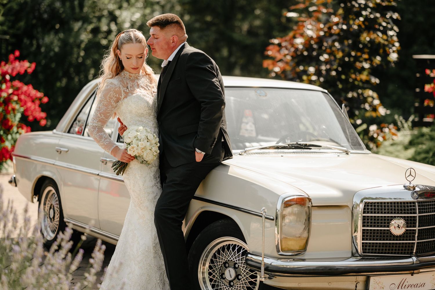 Bride in lace gown and groom in black suit posing beside a classic vintage Mercedes at a wedding.