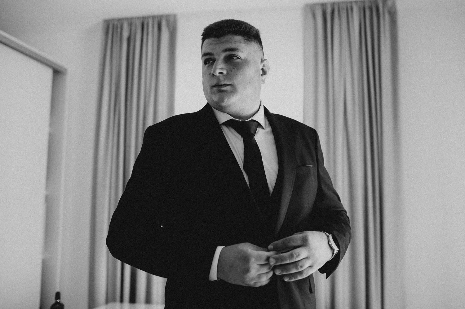 Black and white photo of a man in a dark suit adjusting his jacket button indoors near curtained windows.