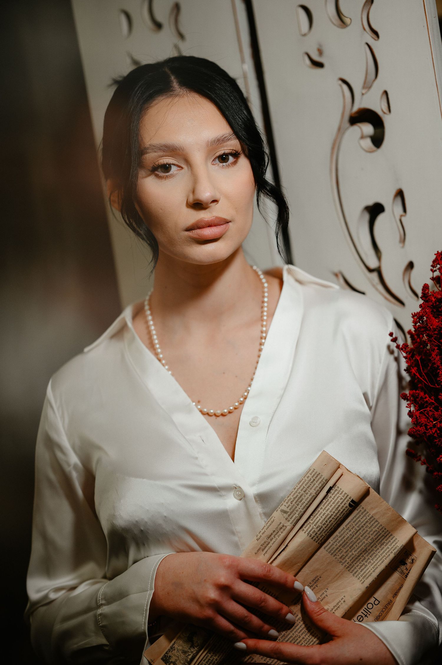 Elegant woman in white silk blouse and pearl necklace holding vintage books, posing near ornate white decor.