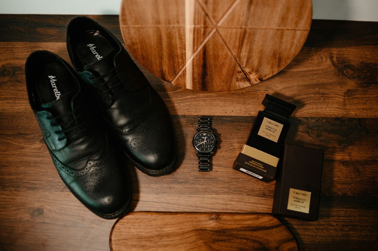 Black leather dress shoes, chronograph watch, and Tom Ford cologne arranged on wooden floor for groom accessories flat lay.