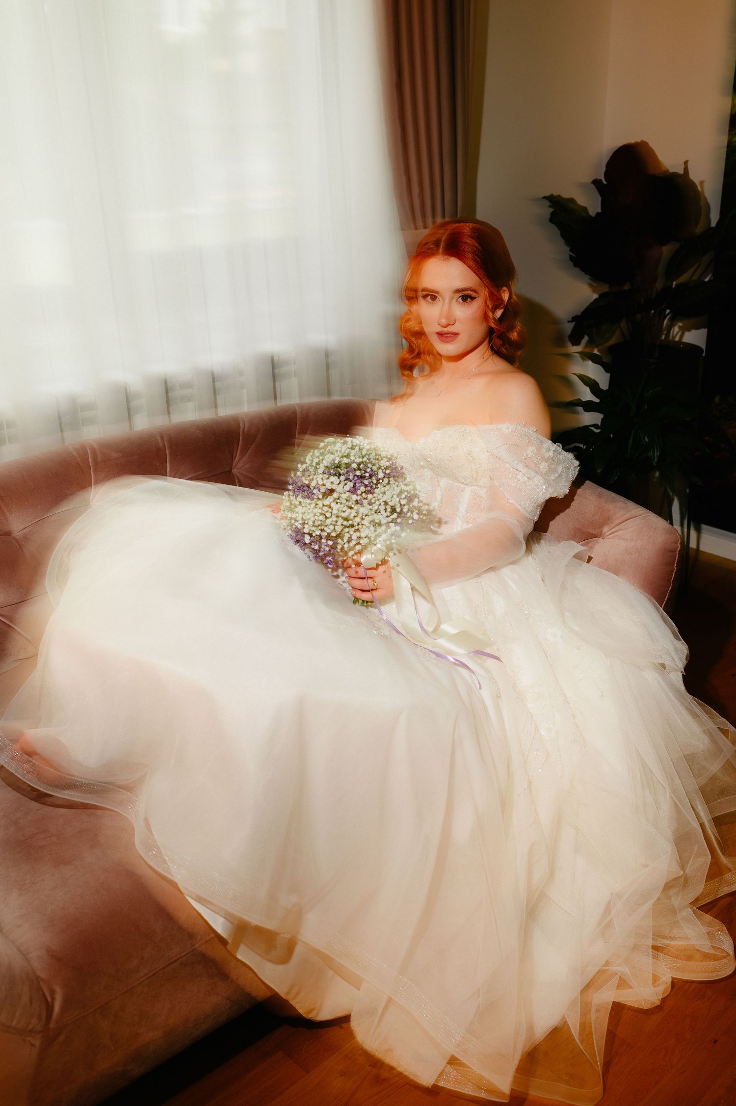 A bride in a white off-shoulder tulle gown sits on a velvet sofa holding a delicate floral bouquet.