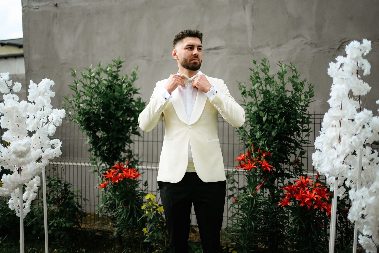 Groom in white tuxedo jacket adjusting bow tie outdoors with floral arrangements.