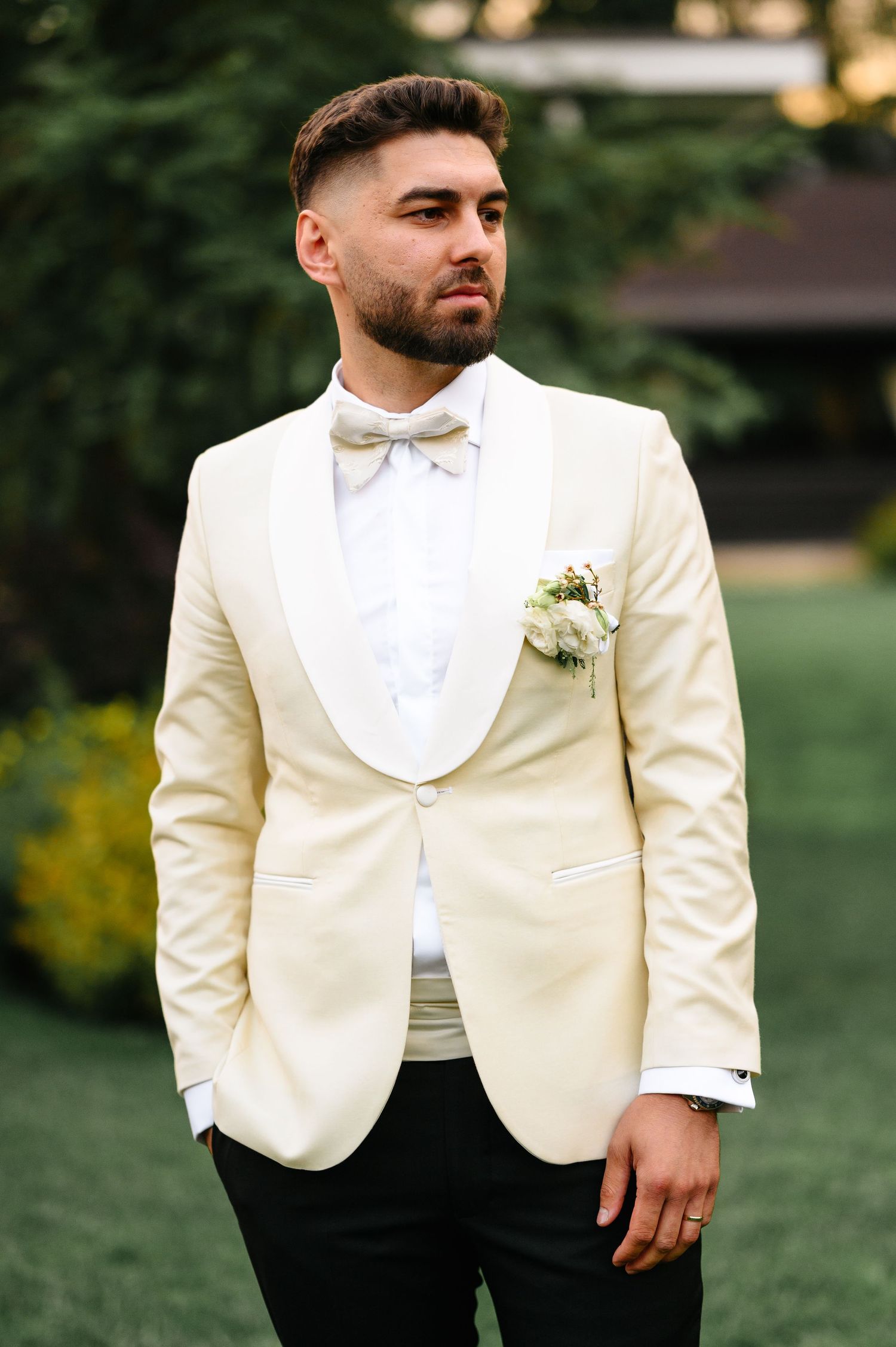 Groom in cream tuxedo jacket with white bow tie and boutonniere posing outdoors at wedding.