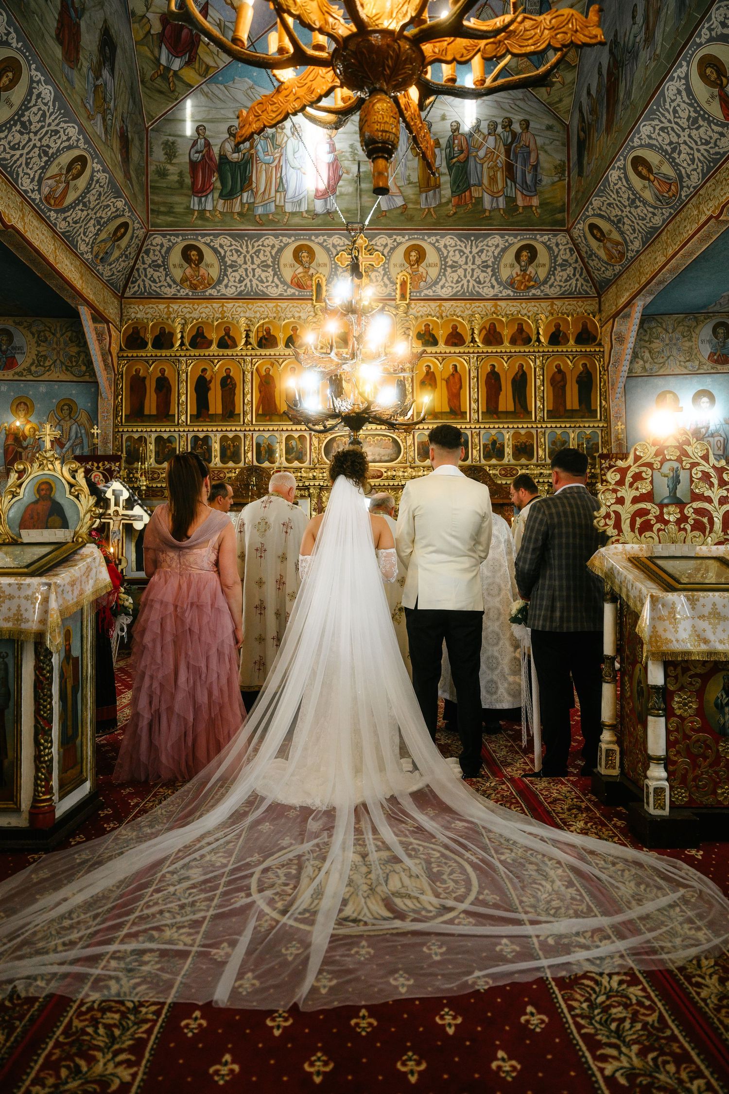 Bride and groom stand at ornate Orthodox church altar during wedding ceremony, bride wearing long cathedral veil.