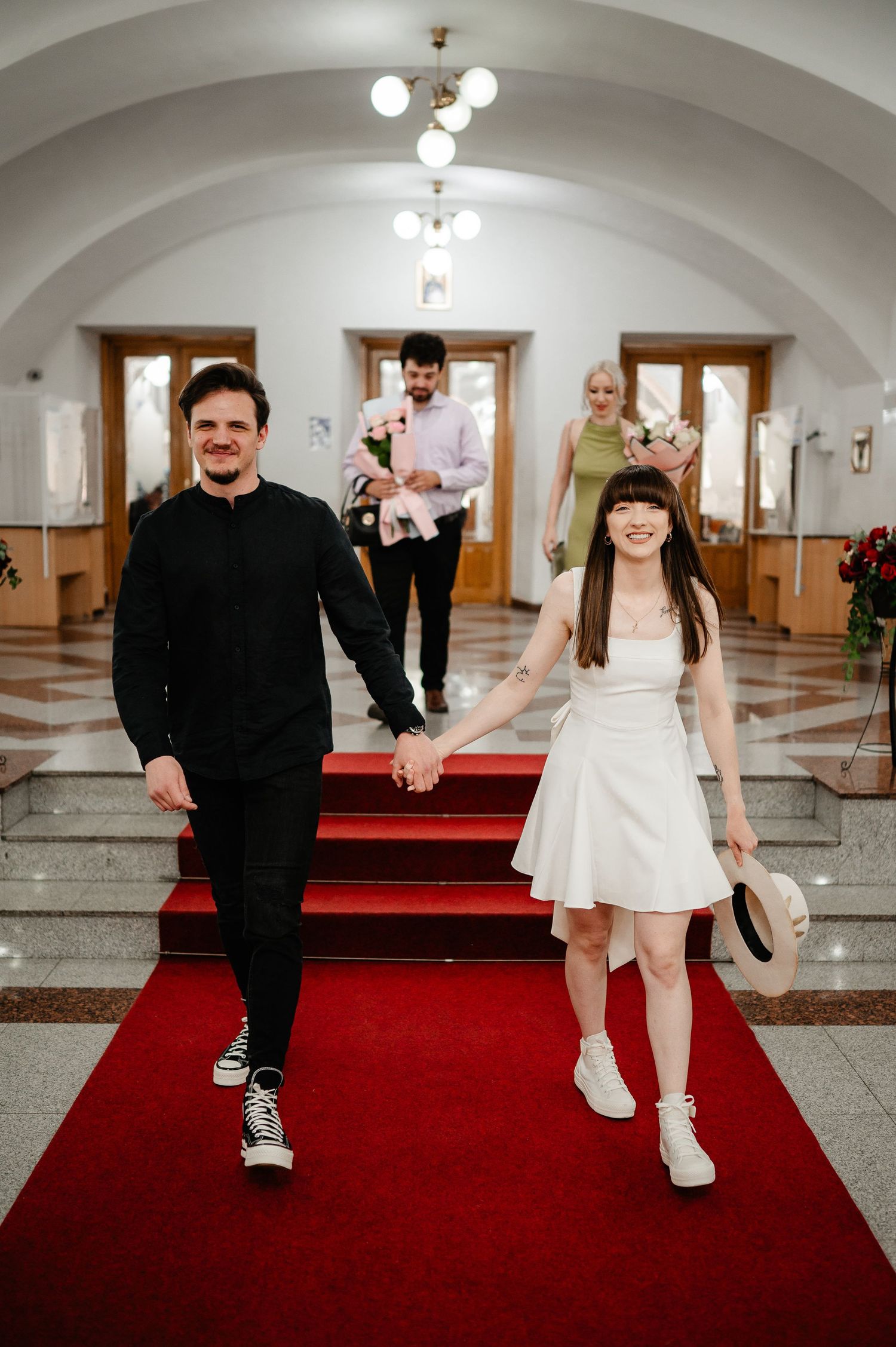 Newlywed couple walks hand-in-hand down red carpet aisle in elegant hall, bride in white mini dress, groom in black.