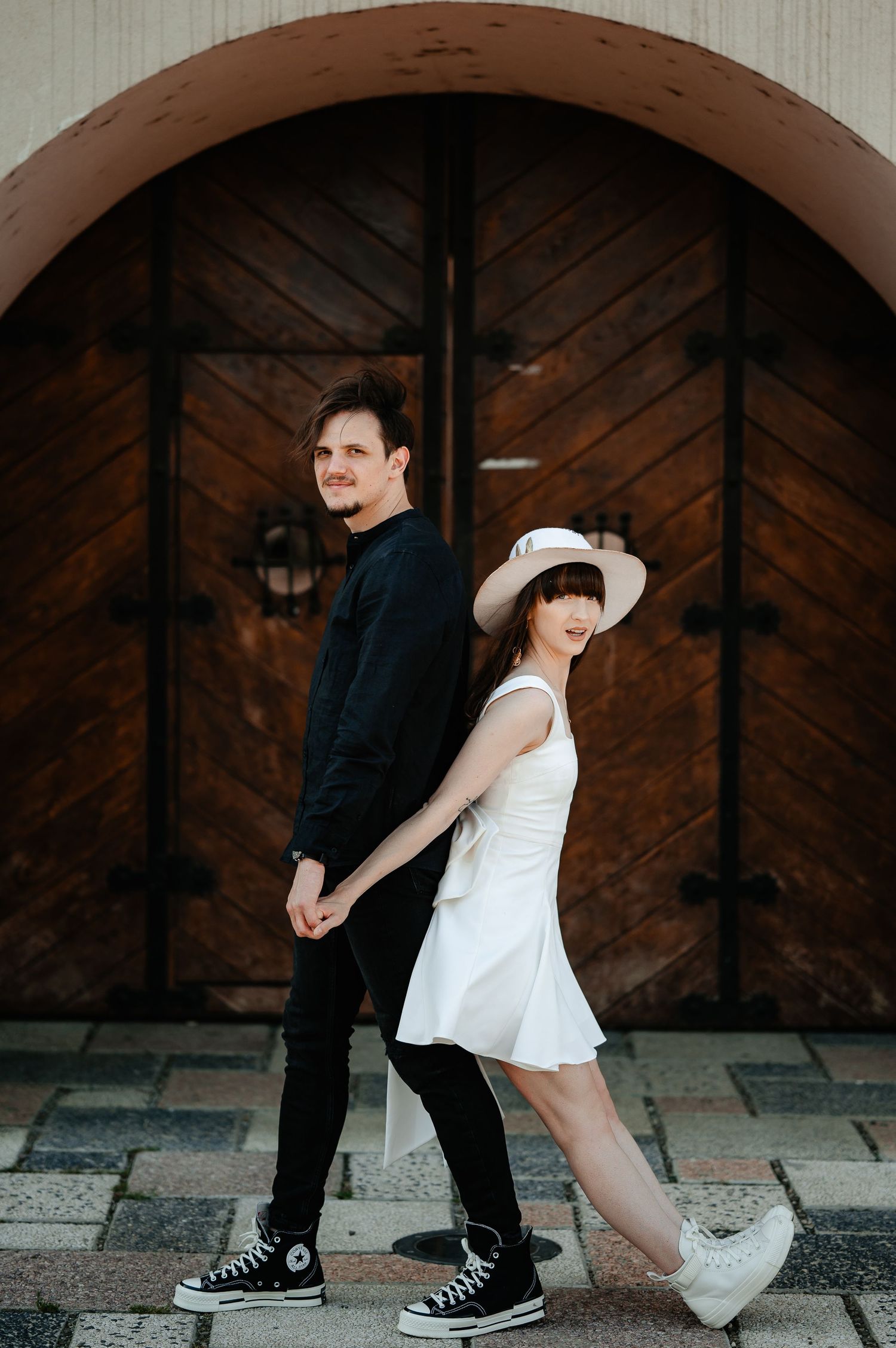 Couple holding hands back-to-back before wooden arched doors, both wearing Converse sneakers.