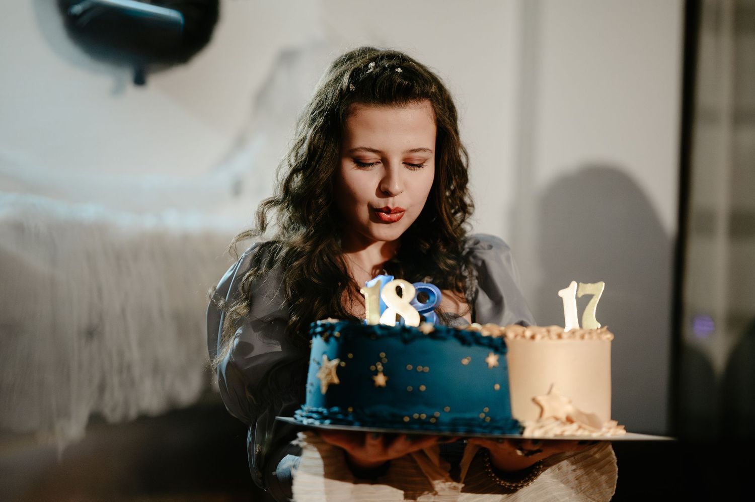 A young woman with curly hair blows out candles on a blue and gold 18th birthday cake.