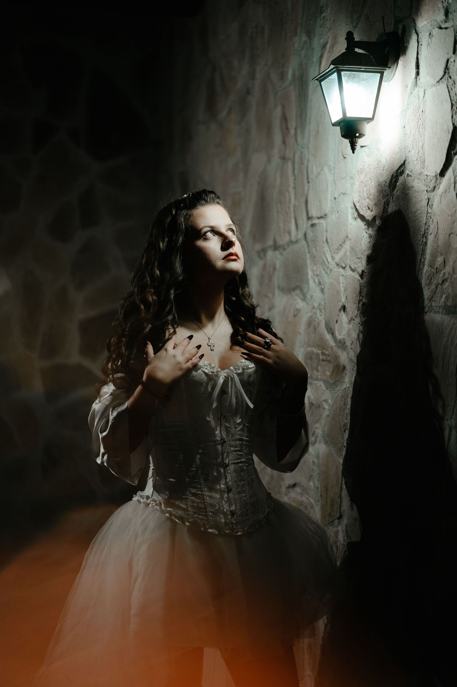 Woman in white corset gown looks up at glowing lantern on stone wall in dark setting.