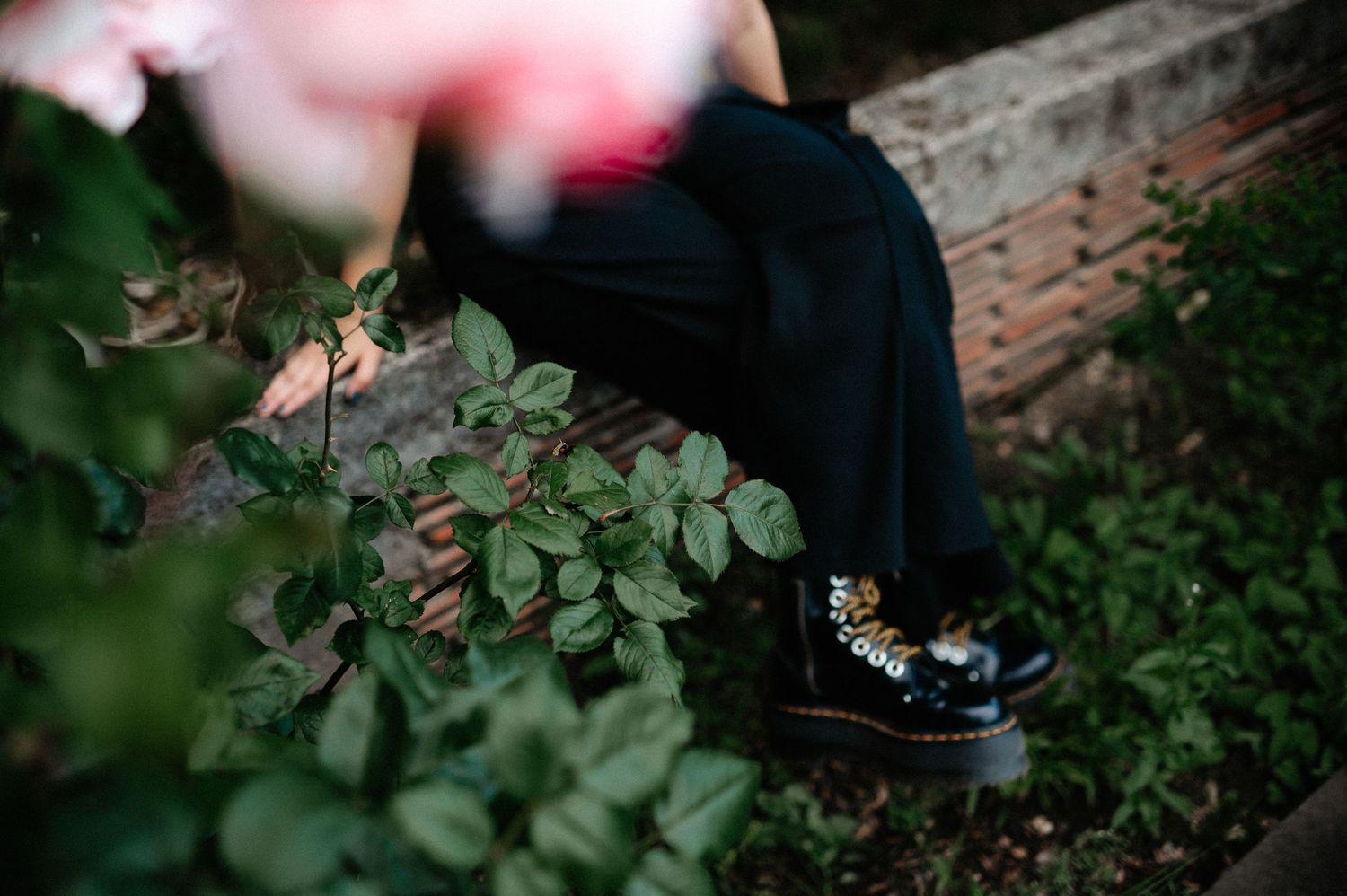Person in black dress and studded boots sits on brick ledge surrounded by green rose bushes.