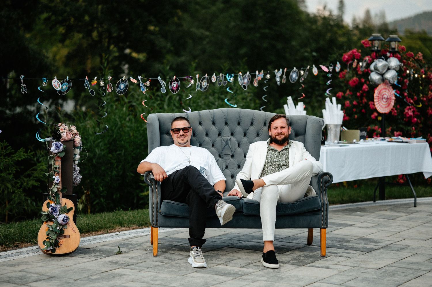 Two men relaxing on a gray tufted sofa at an outdoor garden celebration with guitar nearby.