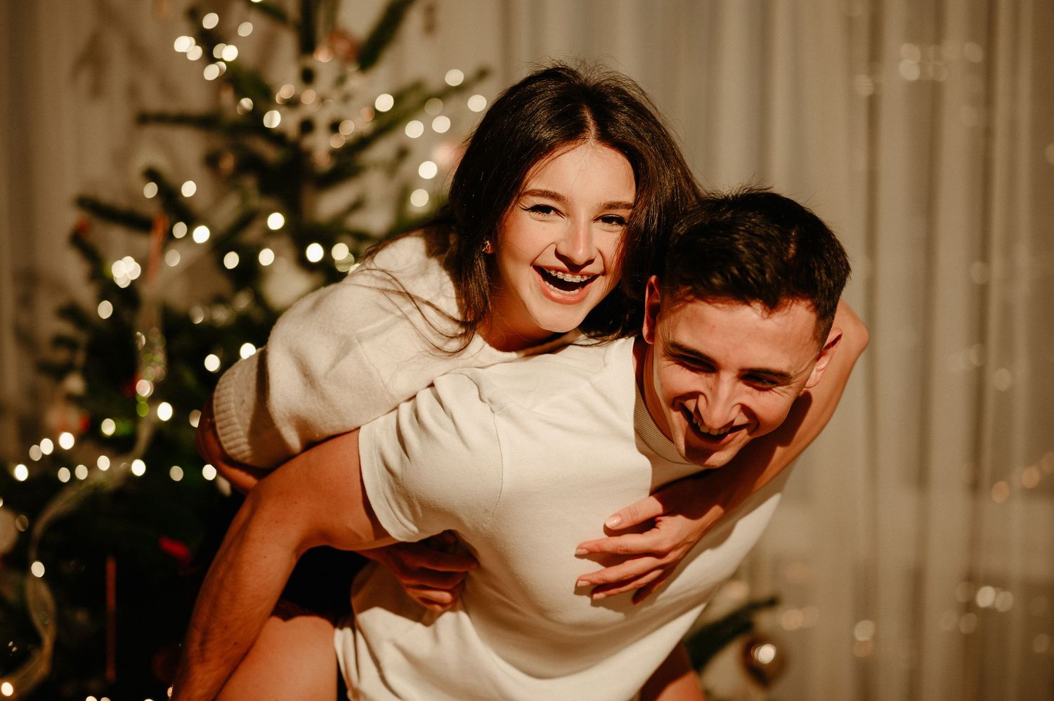 Happy couple sharing a piggyback ride in front of a decorated Christmas tree.