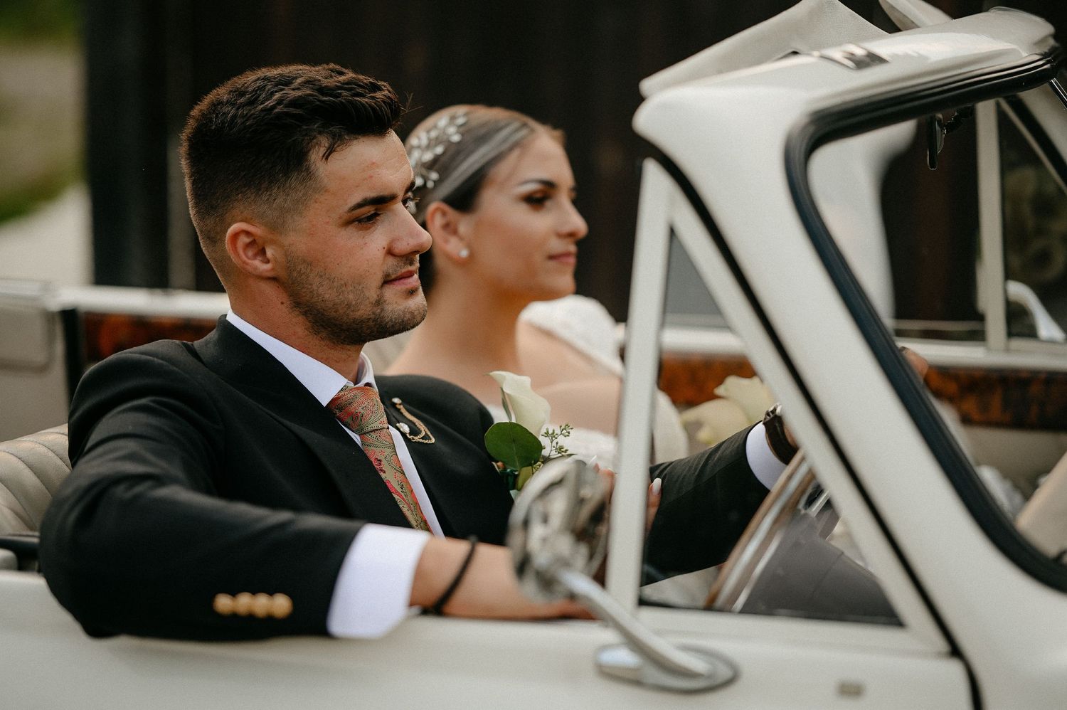 Groom in black suit with floral tie sits in vintage white wedding car with bride in background.