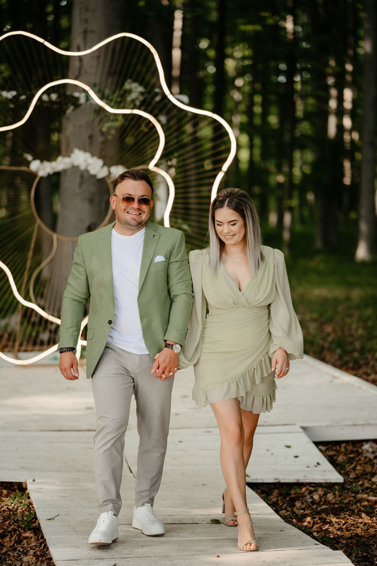Couple in matching sage green outfits walking hand in hand on wooden path with floral neon backdrop.