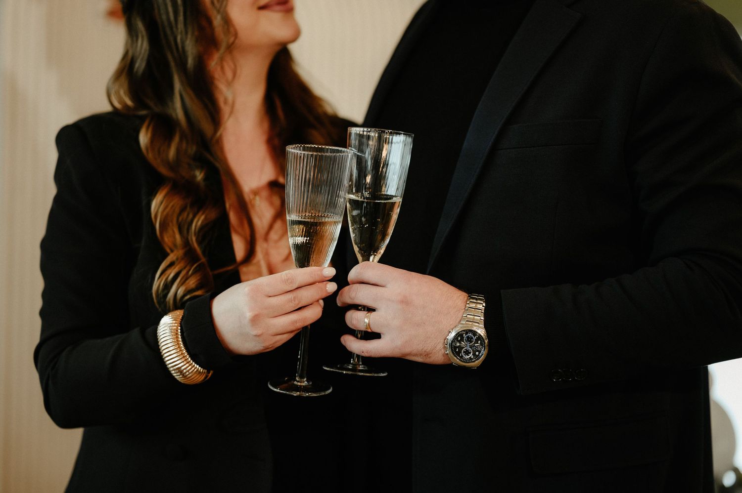 Couple in black attire holding champagne flutes with a luxury watch and gold bracelet visible.