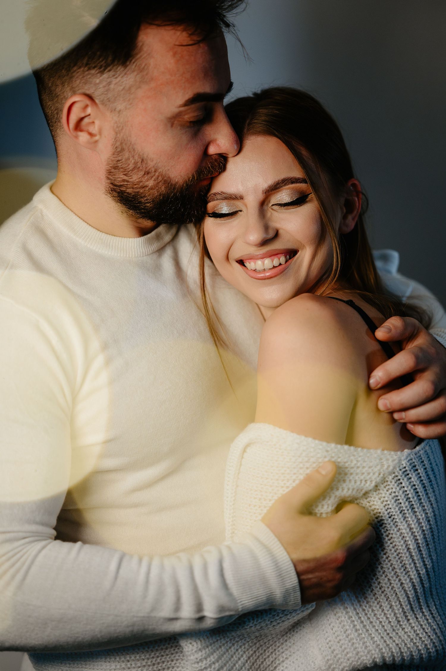 Man in white sweater kisses smiling woman on the cheek in warm sunlit indoor setting.