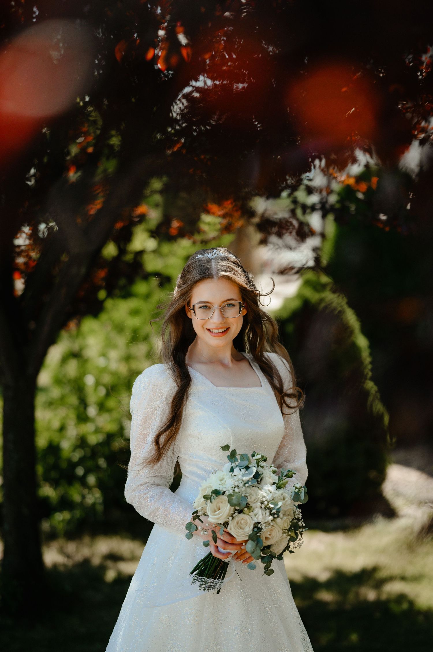 Elegant bride in white lace gown with glasses holding bouquet in sunny garden.