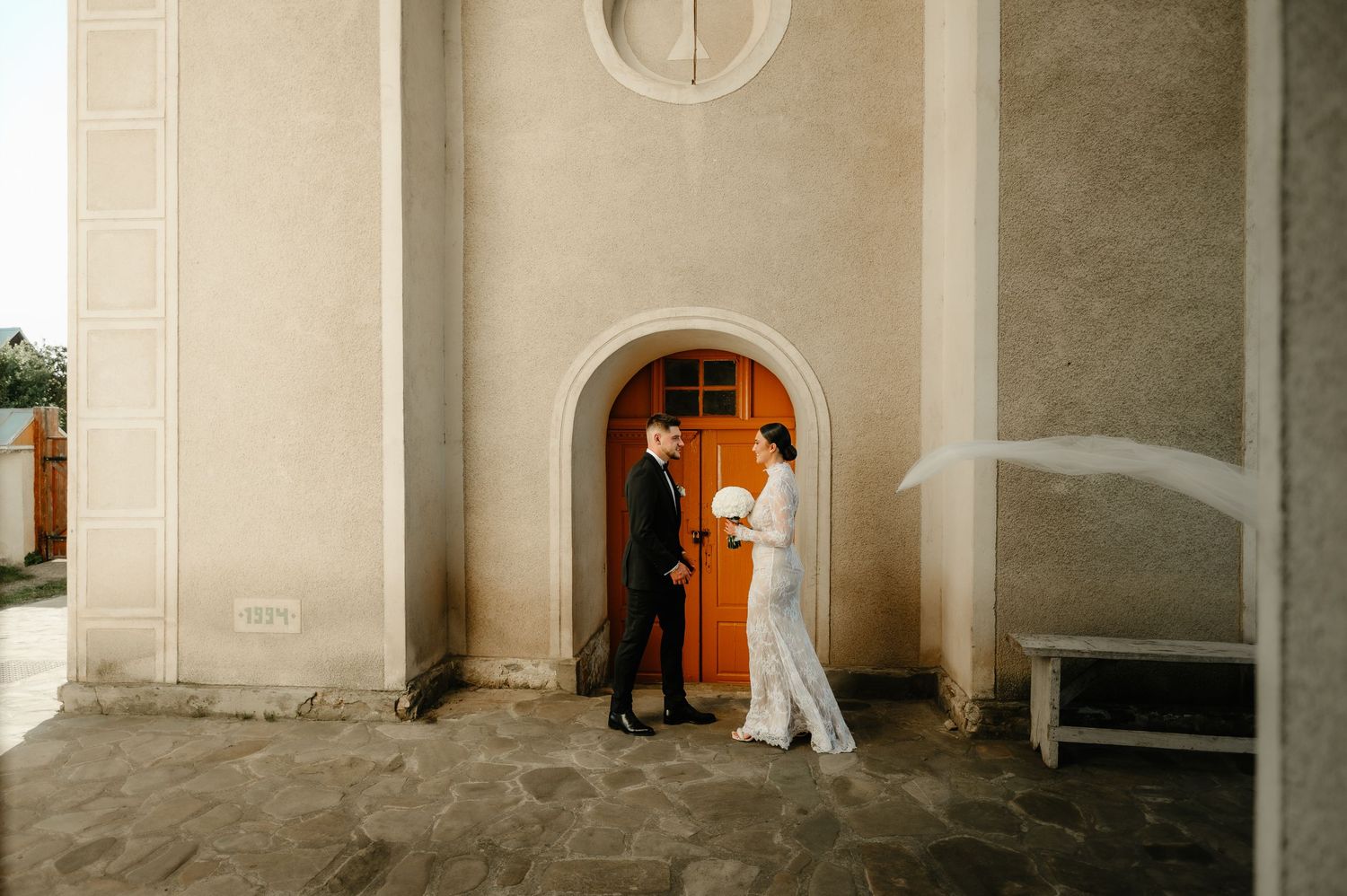 Bride and groom standing outside a church entrance with an orange arched door on their wedding day.