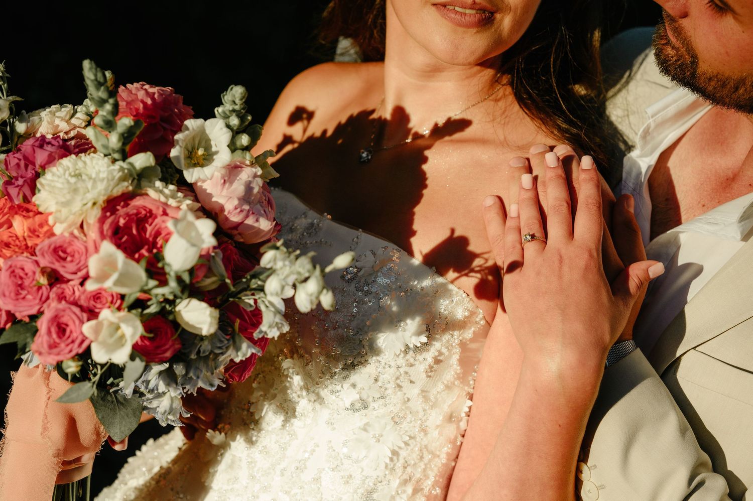 Bride in lace wedding dress holding pink and white bouquet with groom, showing diamond engagement ring.