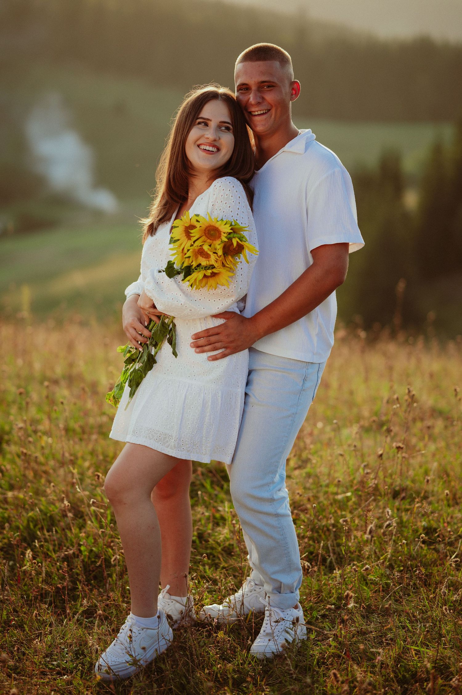 Happy couple in white outfits embracing with sunflowers in a sunlit mountain field.