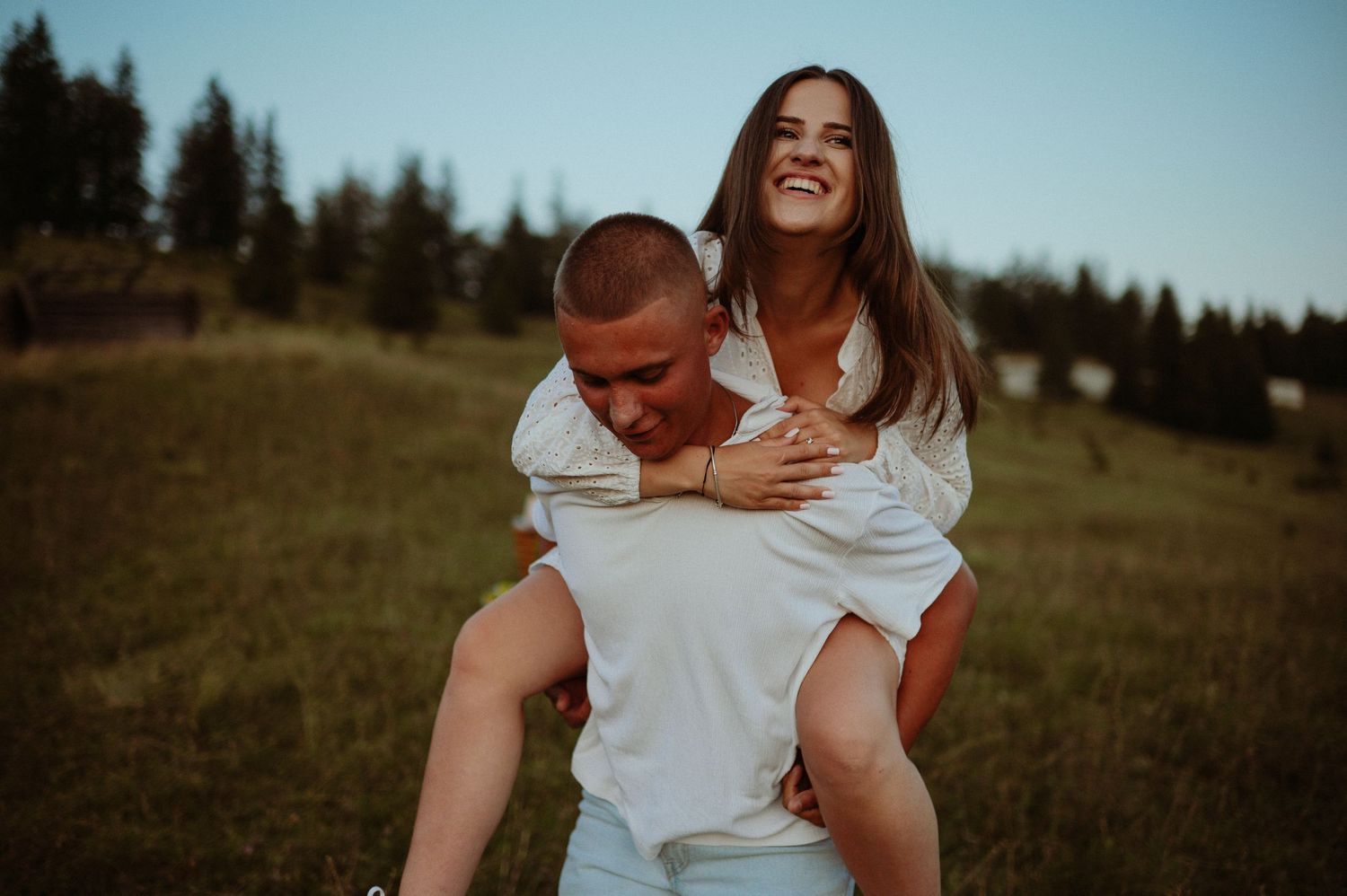 Joyful woman laughing while getting a piggyback ride from man in white shirt on green hillside at sunset.
