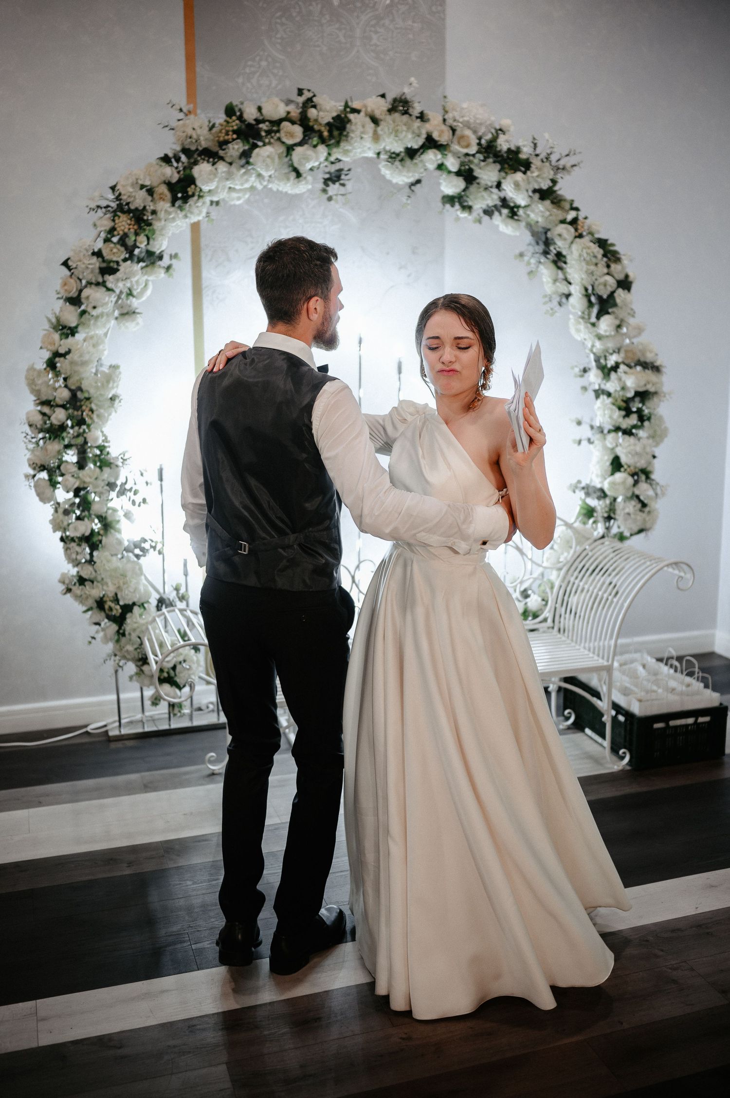 Bride in elegant white gown reading vows near a stunning circular white rose floral arch.