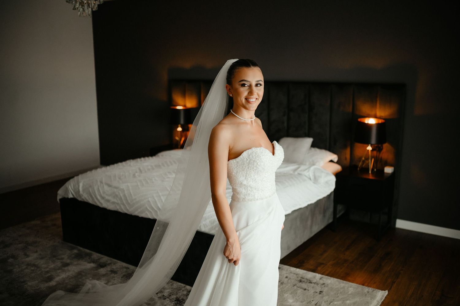 Smiling bride in strapless white gown holding veil in warmly lit bedroom.