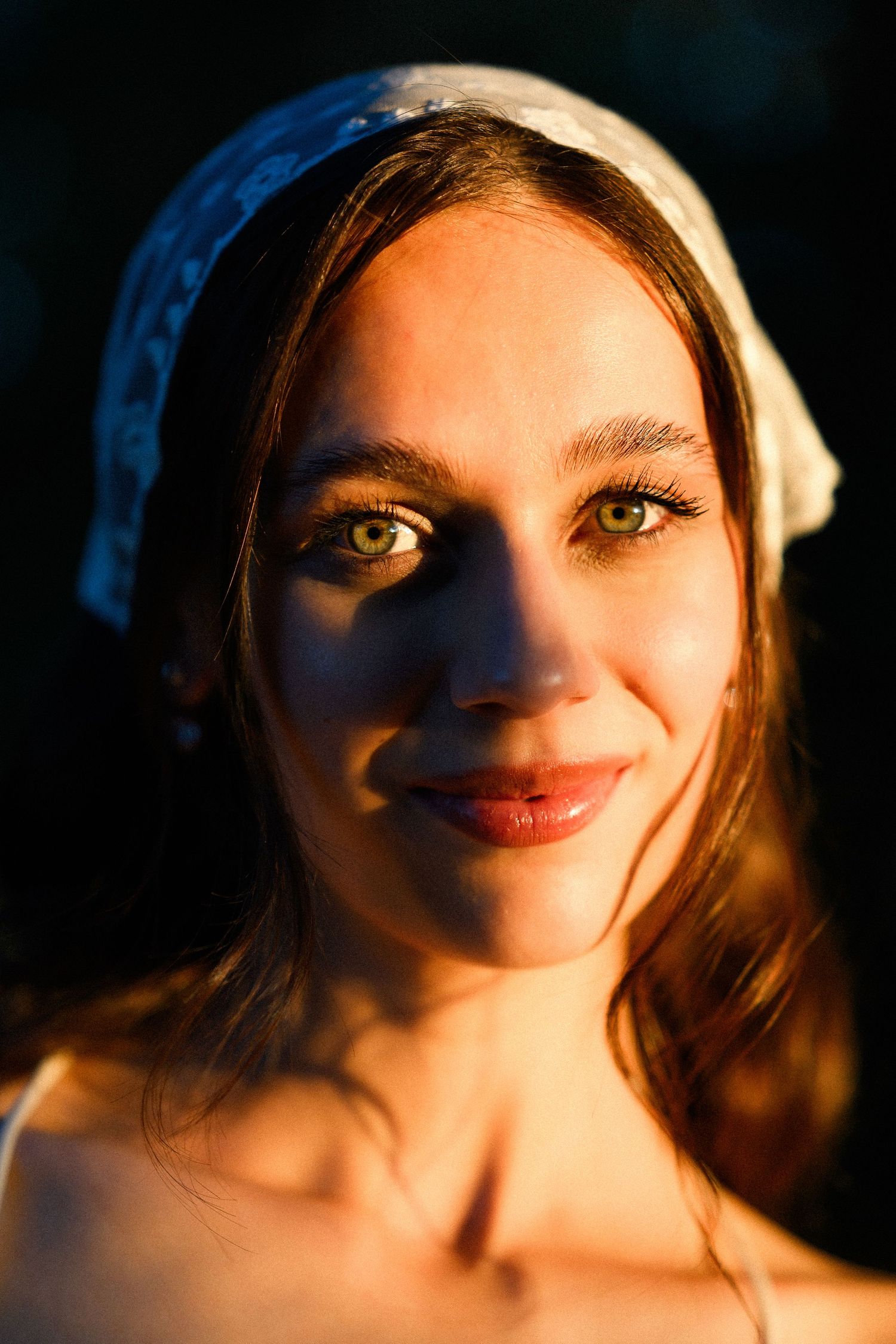 Young woman with green eyes and white headband bathed in warm golden hour light against dark background.