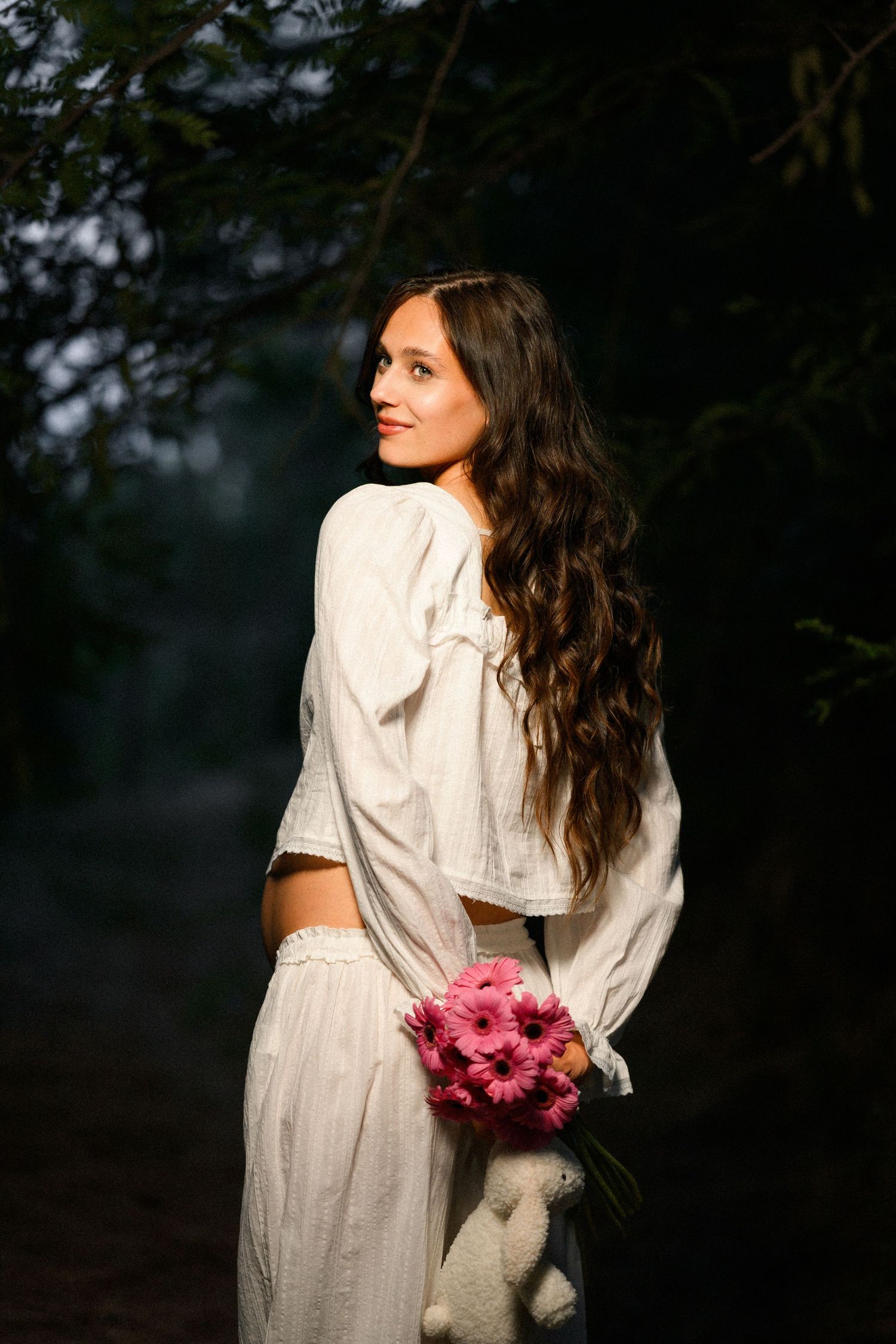 Young woman in white outfit holds pink gerbera bouquet, smiling over shoulder in misty forest setting.