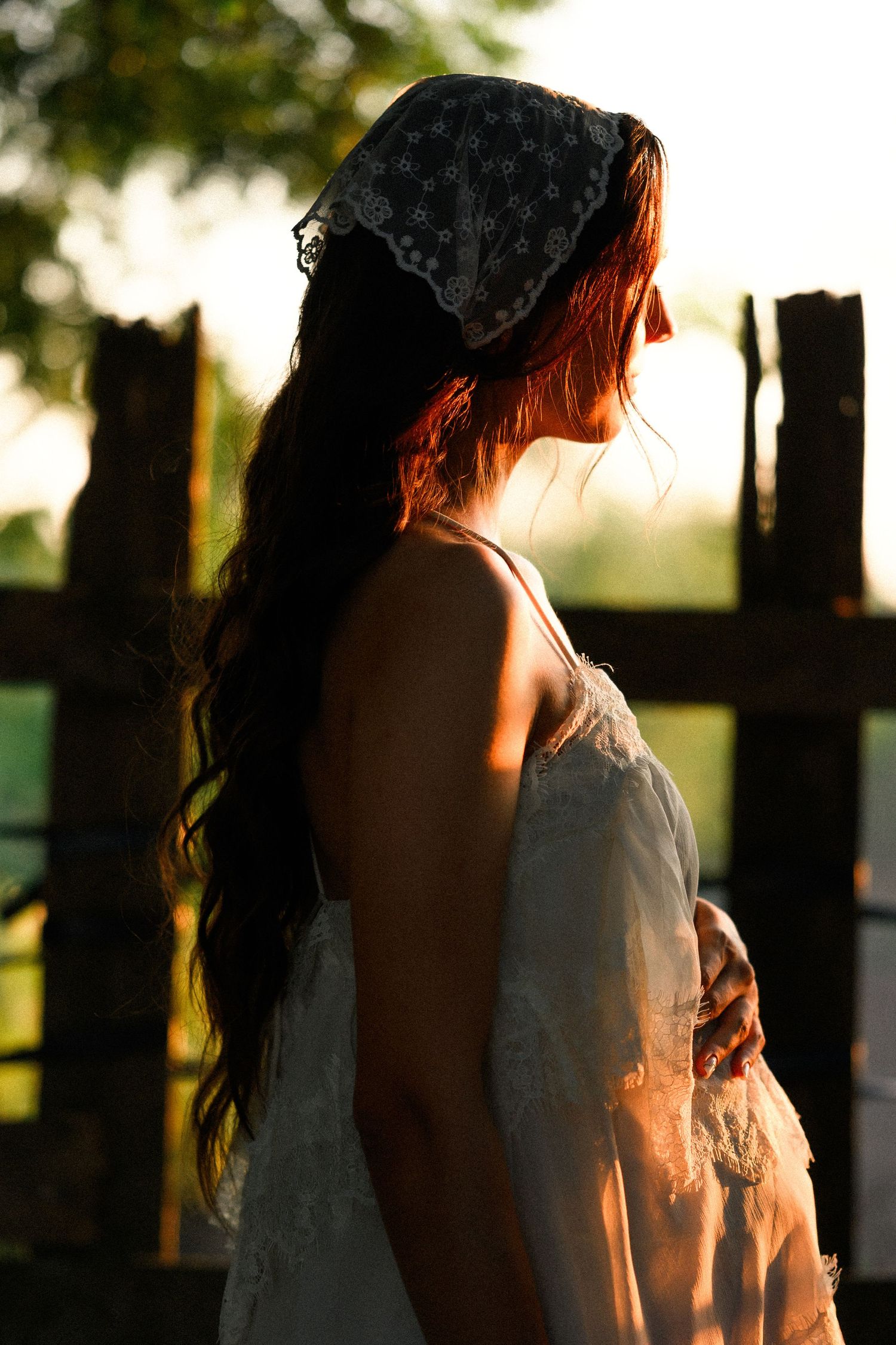 Woman in white dress and lace headscarf with long dark hair stands in golden sunset light near wooden fence.