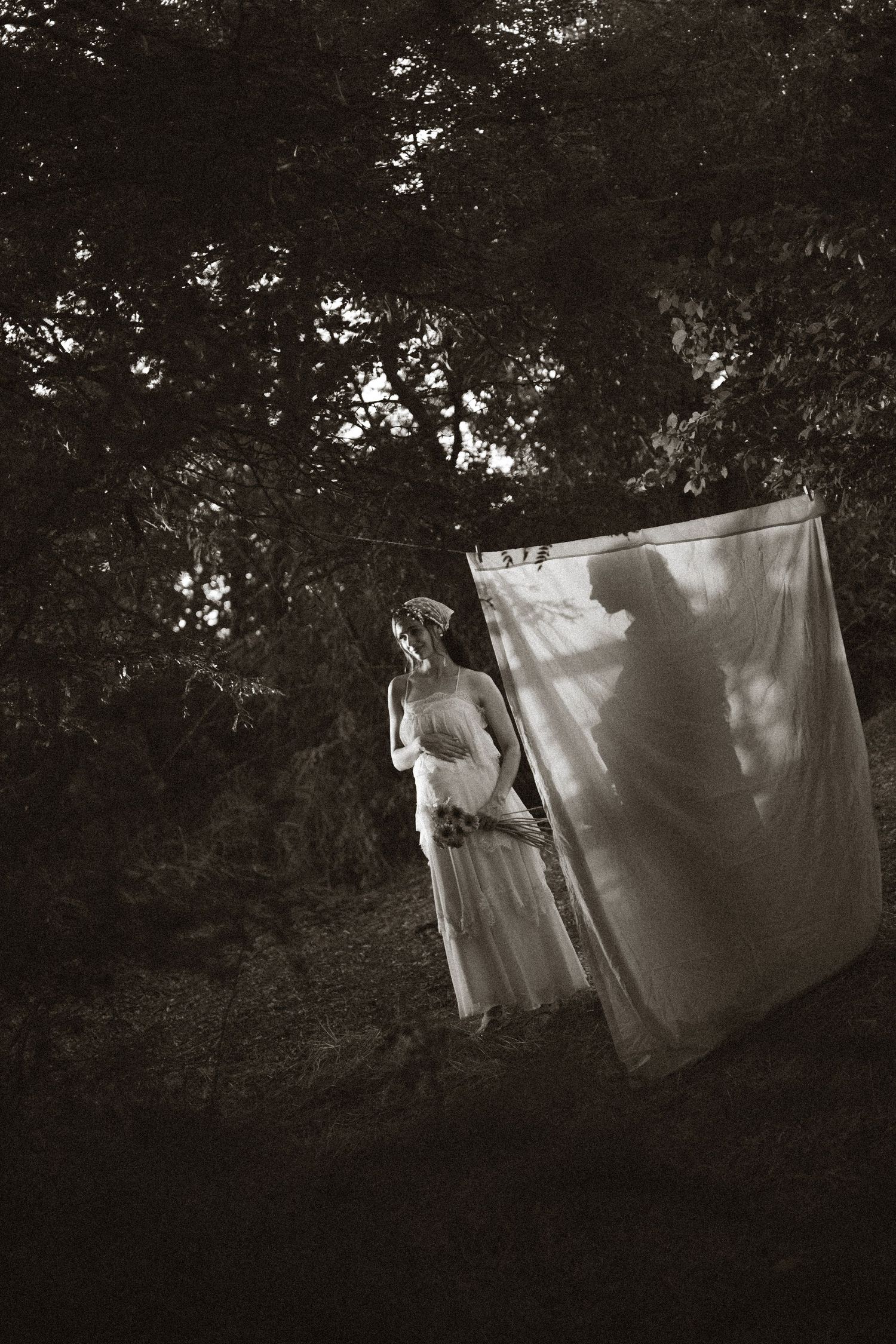 Black and white photo of a woman in white dress standing by hanging sheet in dark forest.