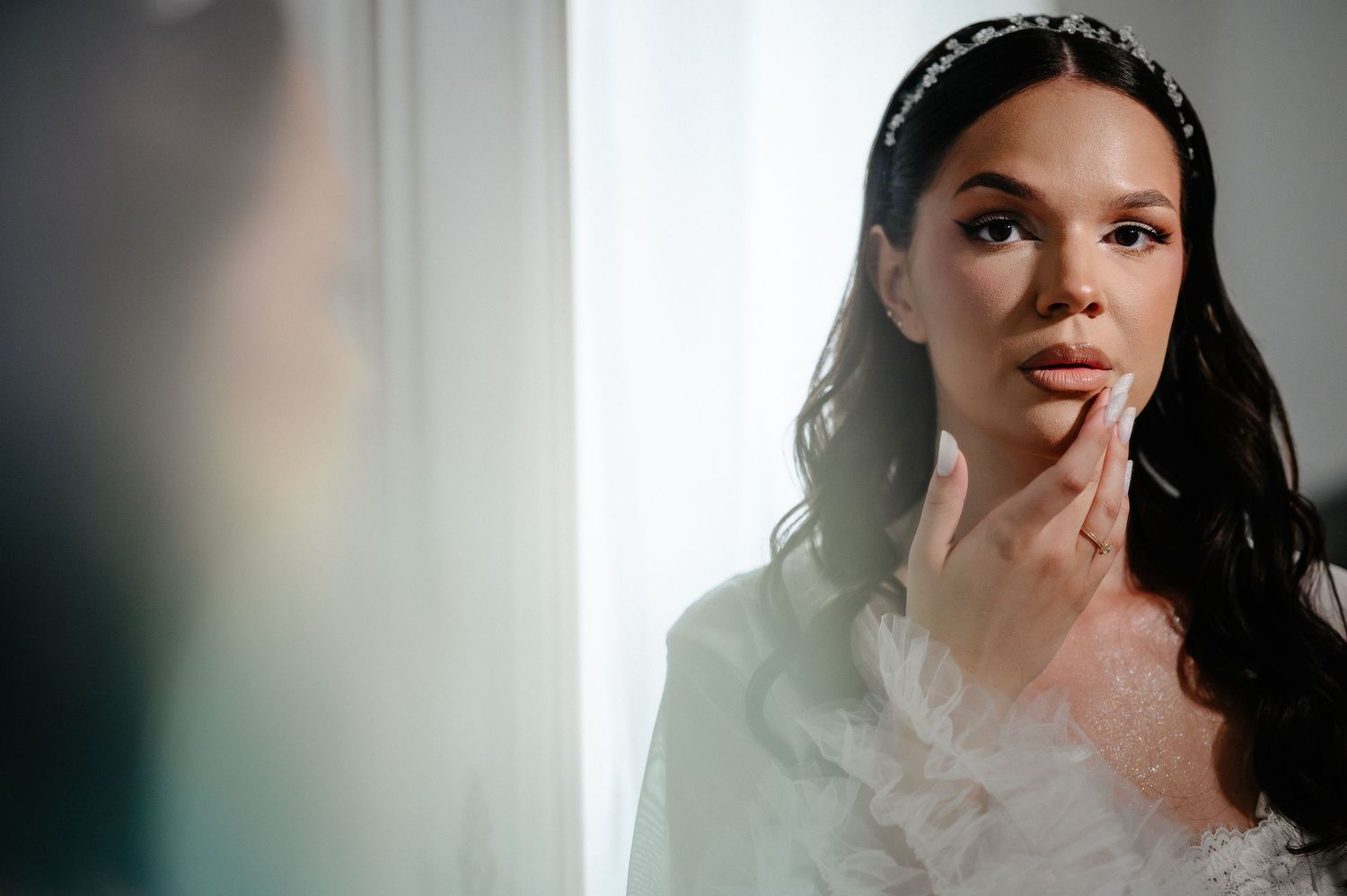Bride in white robe touching her face while gazing into mirror during wedding morning preparation.