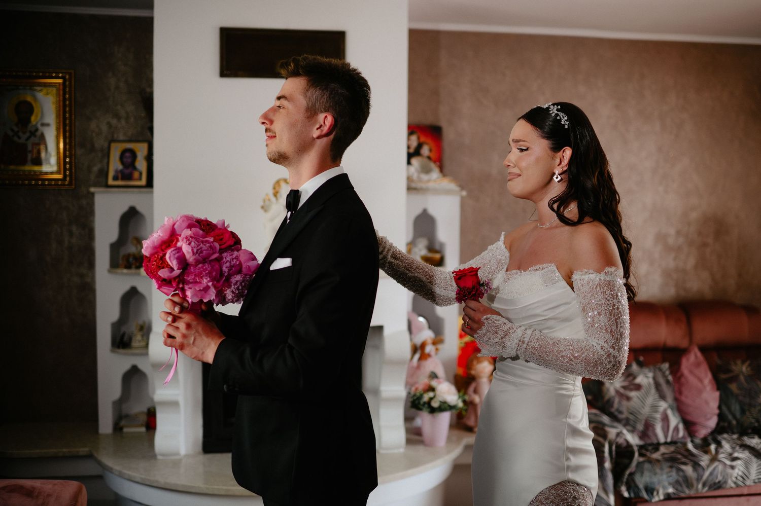 Groom in black suit and bride in white dress sharing first look moment with pink peonies.
