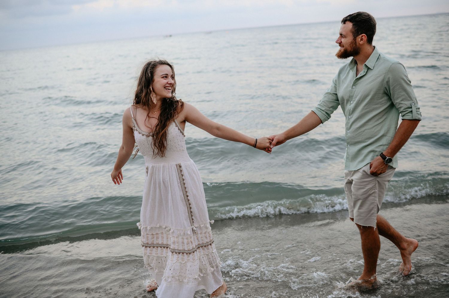 Joyful couple holding hands and laughing while walking through gentle ocean waves on a beach.
