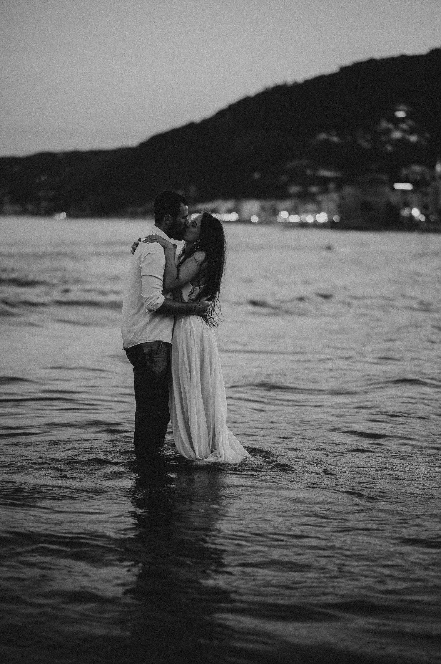 Black and white photo of a couple embracing and kissing while standing in ocean water at dusk.