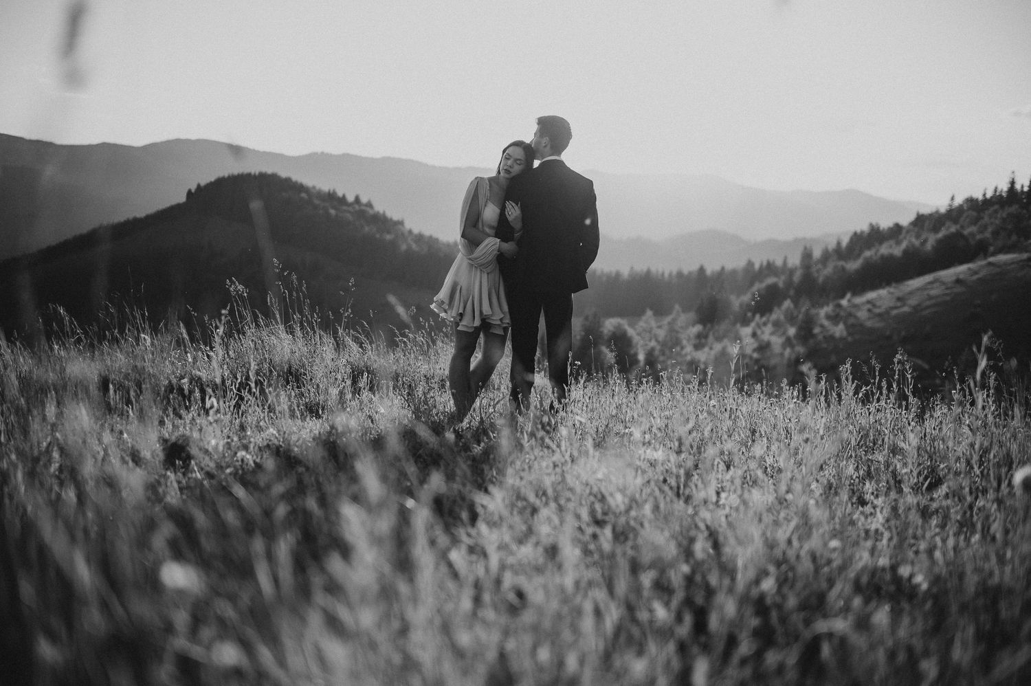 Black and white photo of couple embracing on a grassy mountain hillside at sunset.