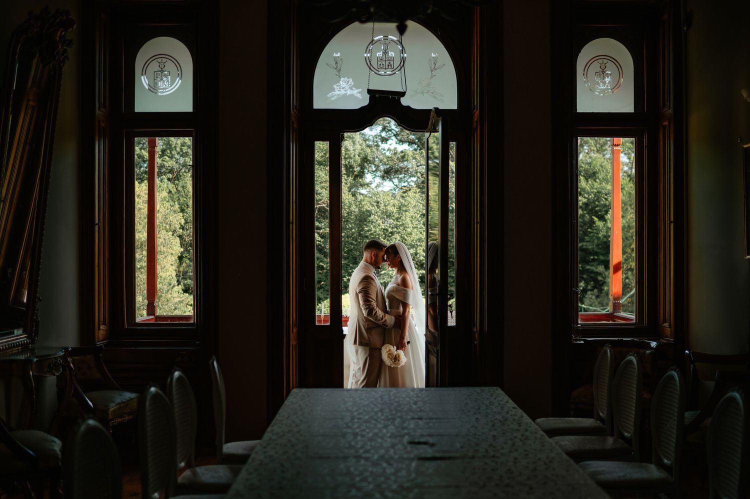 Romantic silhouette of bride and groom kissing in ornate arched doorway with lush green garden visible behind them.