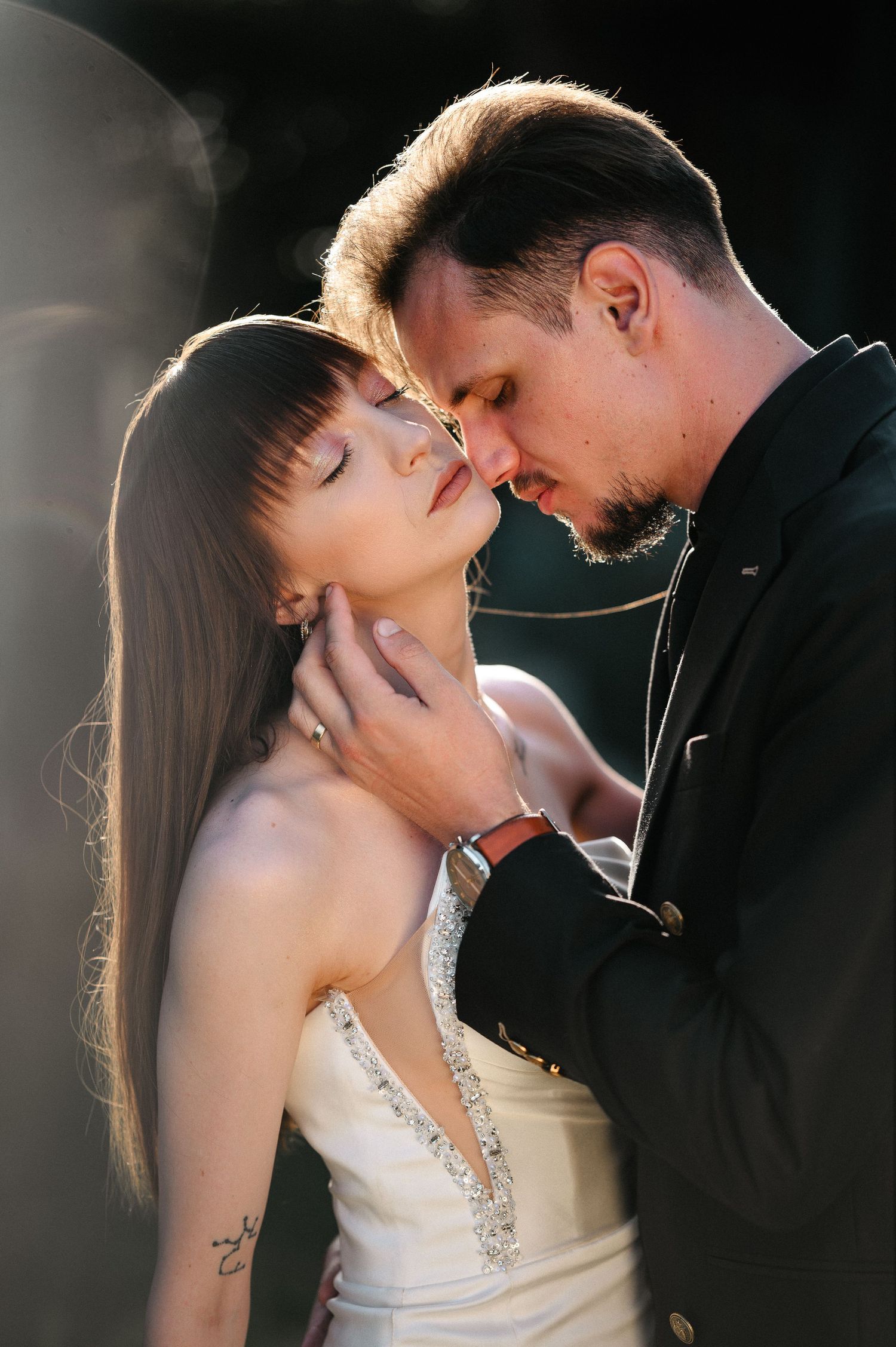 Groom tenderly holds bride's face as they share an intimate almost-kiss in golden backlit wedding portrait.