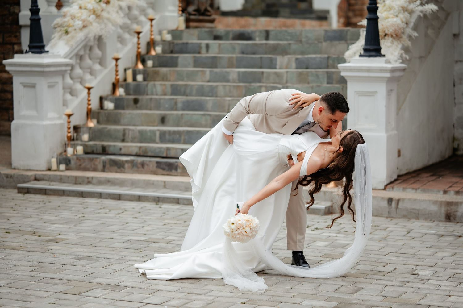 Groom dramatically dips bride in romantic dance pose on cobblestone courtyard in front of white staircase wedding venue.