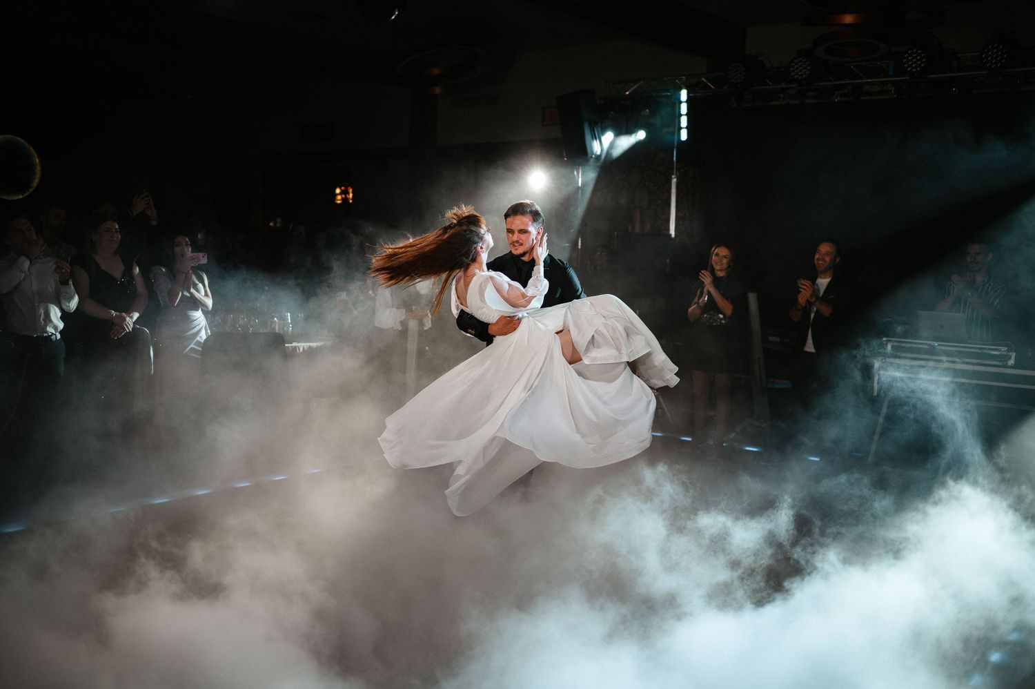 Groom lifts bride in flowing white dress during energetic first dance surrounded by wedding fog effects.
