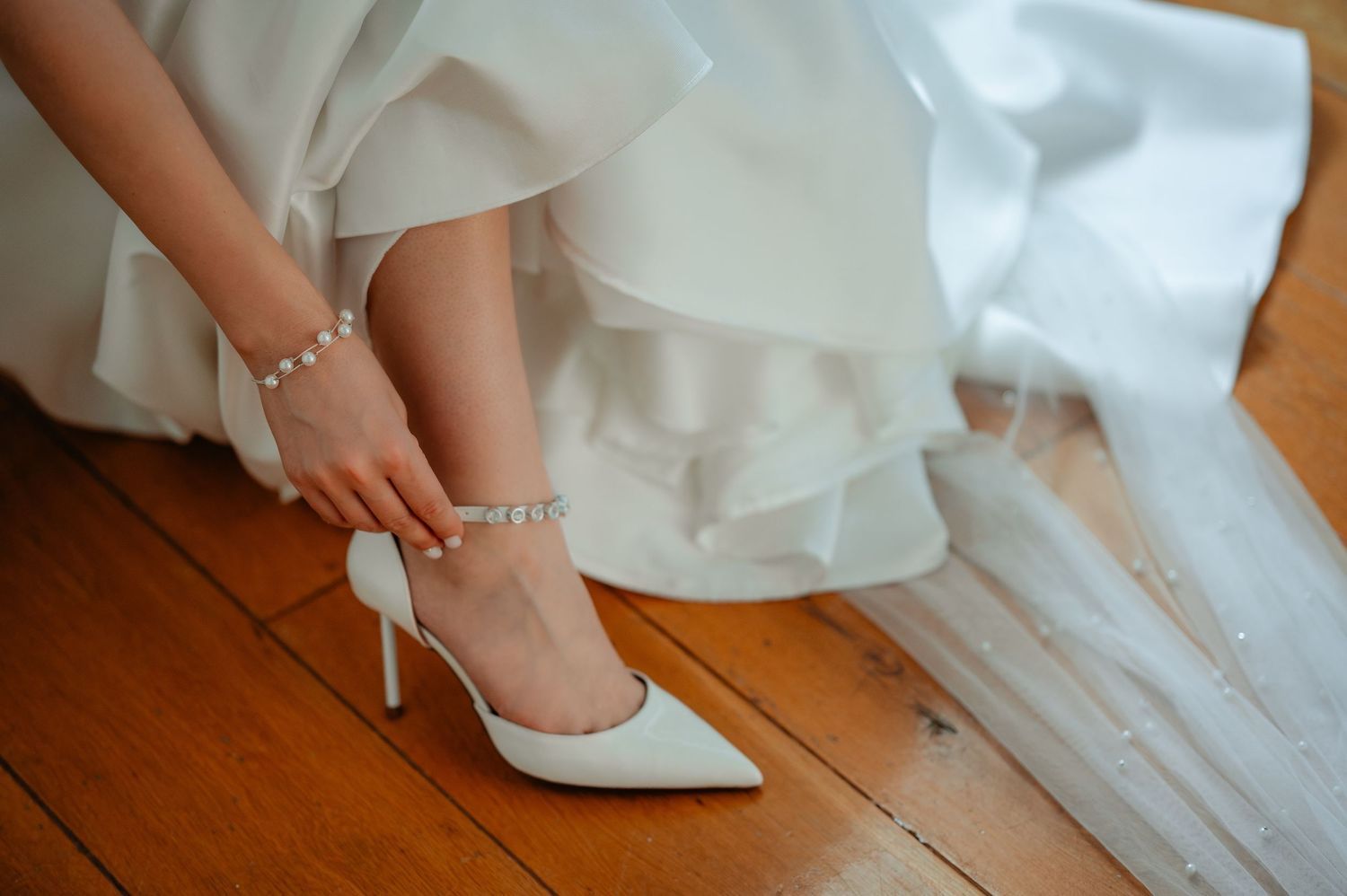 Bride fastening a pearl anklet on her foot while wearing white pointed-toe heels on a wood floor.