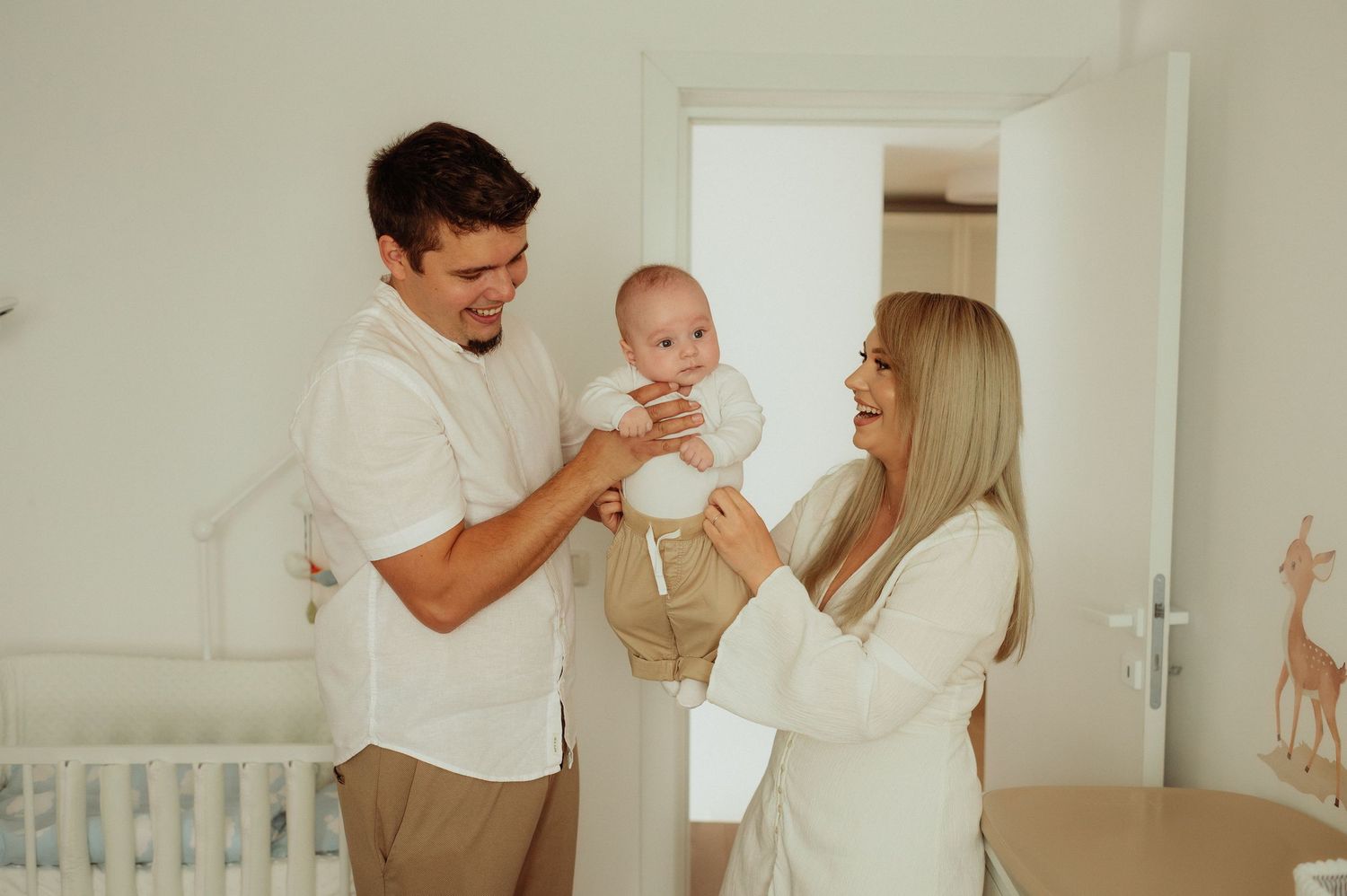 Happy young parents in white clothing playing with baby in bright nursery.