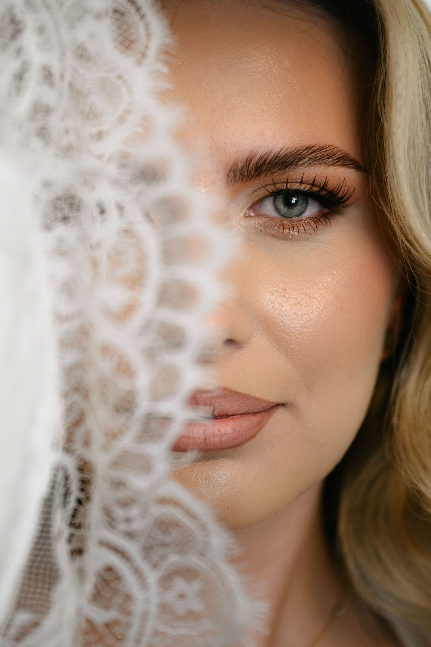 Close-up portrait of a bride with elegant makeup, half her face covered by a delicate white lace wedding veil.