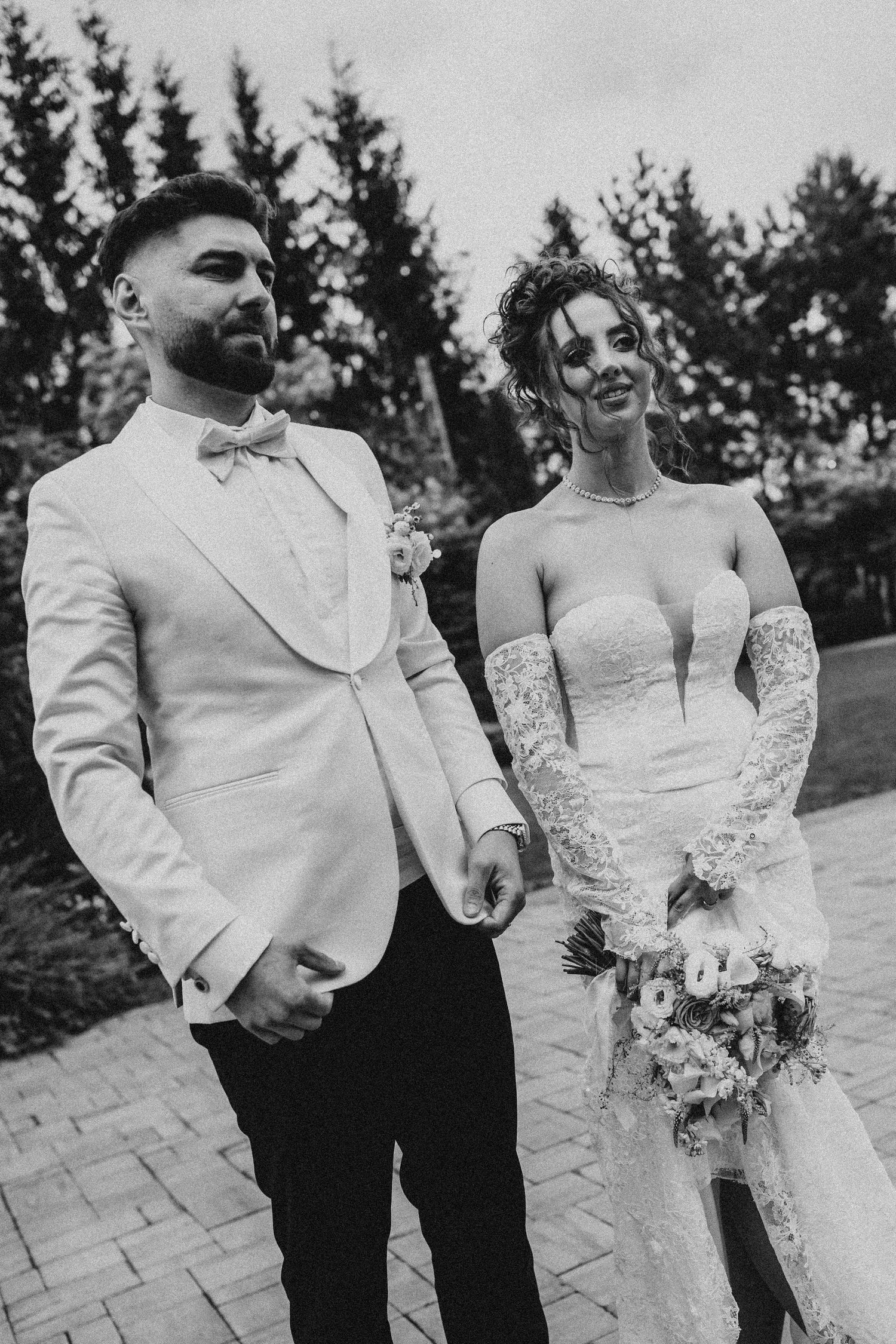 Black and white photo of a bride in lace gown holding bouquet and groom in white tuxedo jacket on wedding day.